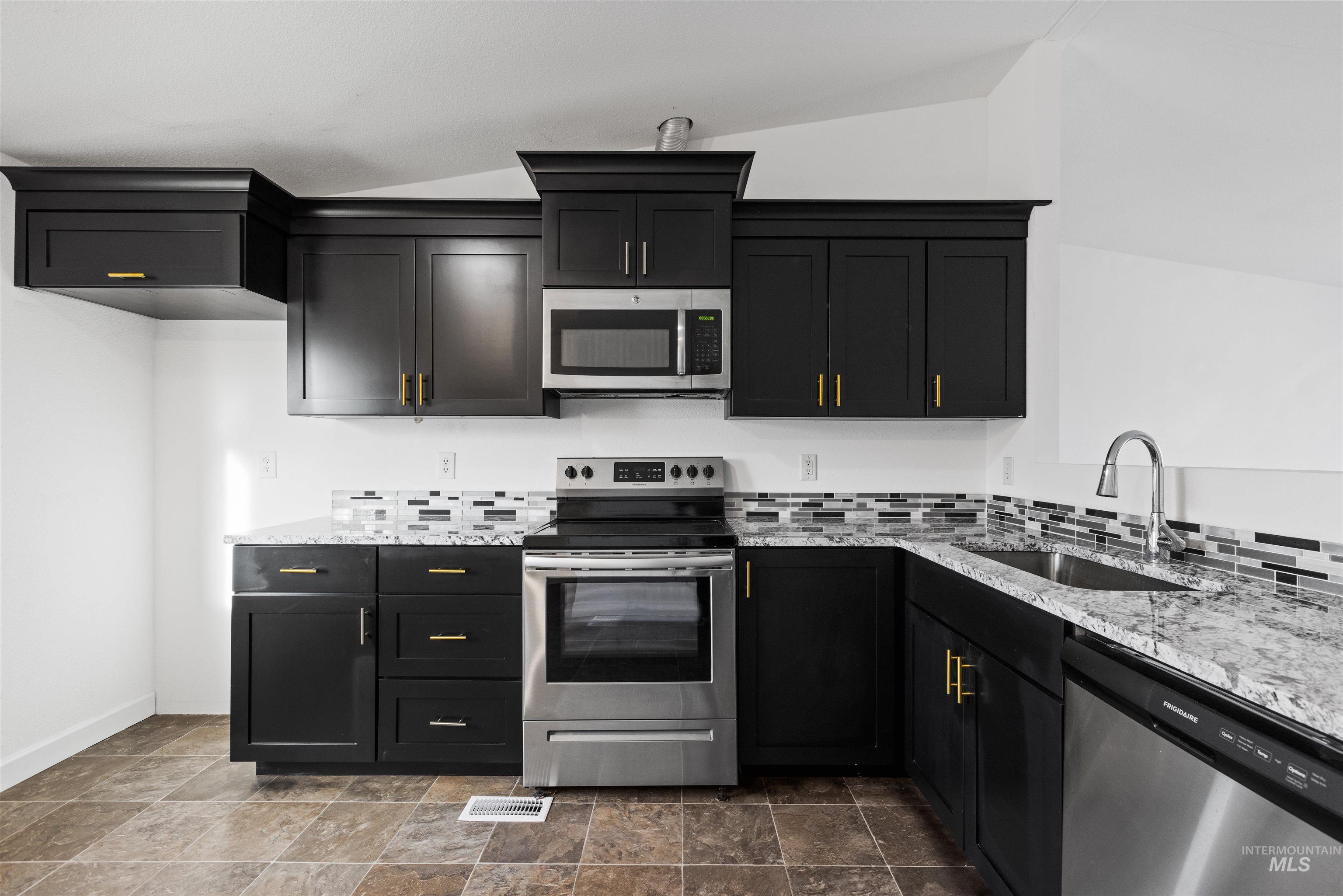 Kitchen with stainless steel appliances, light stone countertops, dark cabinetry, vaulted ceiling, and stone finish flooring