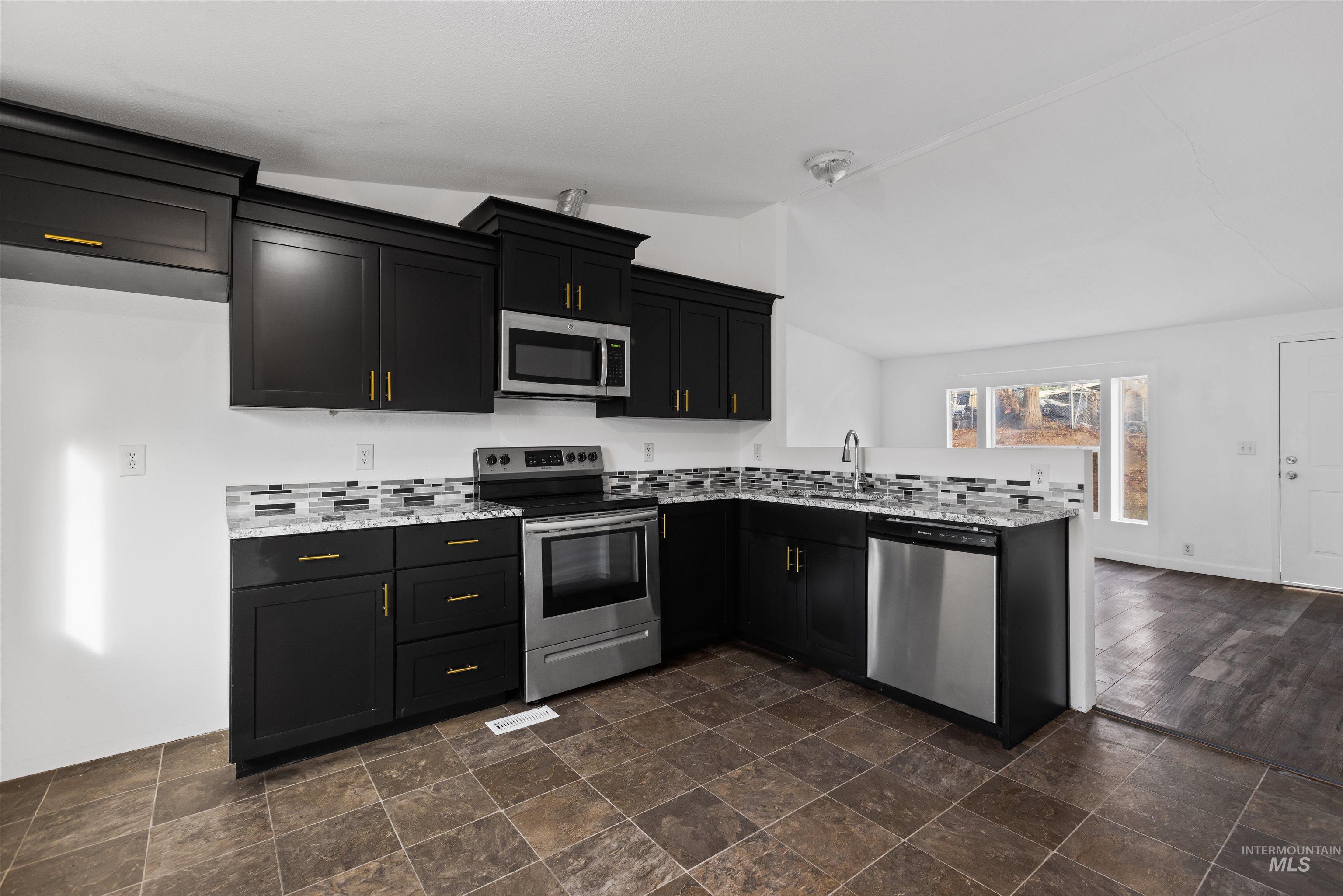 Kitchen featuring vaulted ceiling, appliances with stainless steel finishes, a peninsula, dark cabinetry, and stone finish flooring