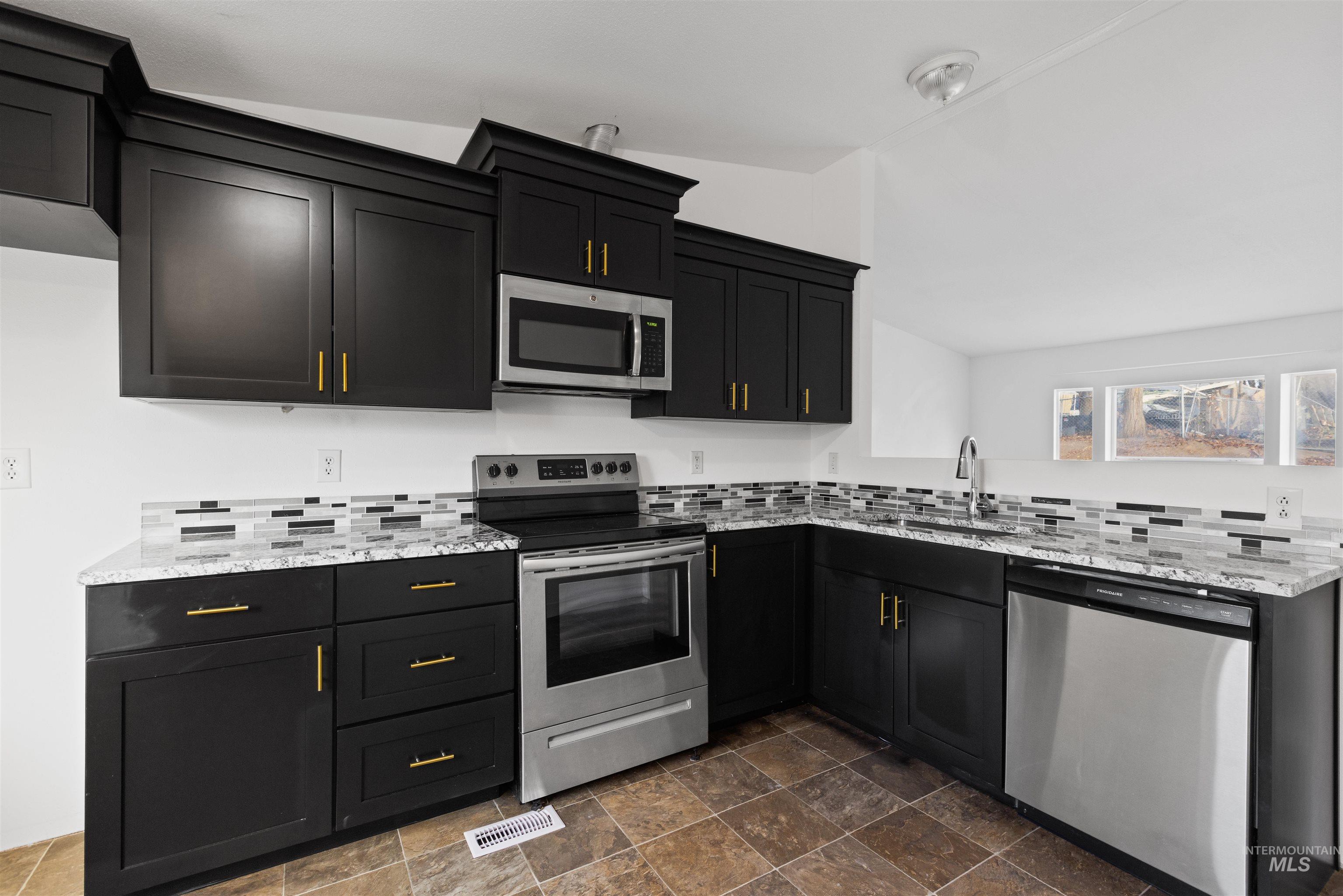 Kitchen with appliances with stainless steel finishes, light stone counters, dark cabinets, stone finish floors, and vaulted ceiling