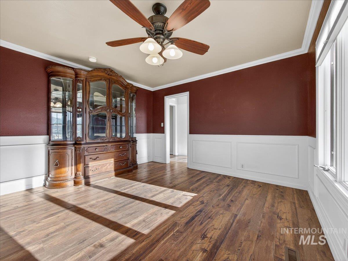 Empty room with a wainscoted wall, dark wood-style flooring, crown molding, ceiling fan, and a decorative wall
