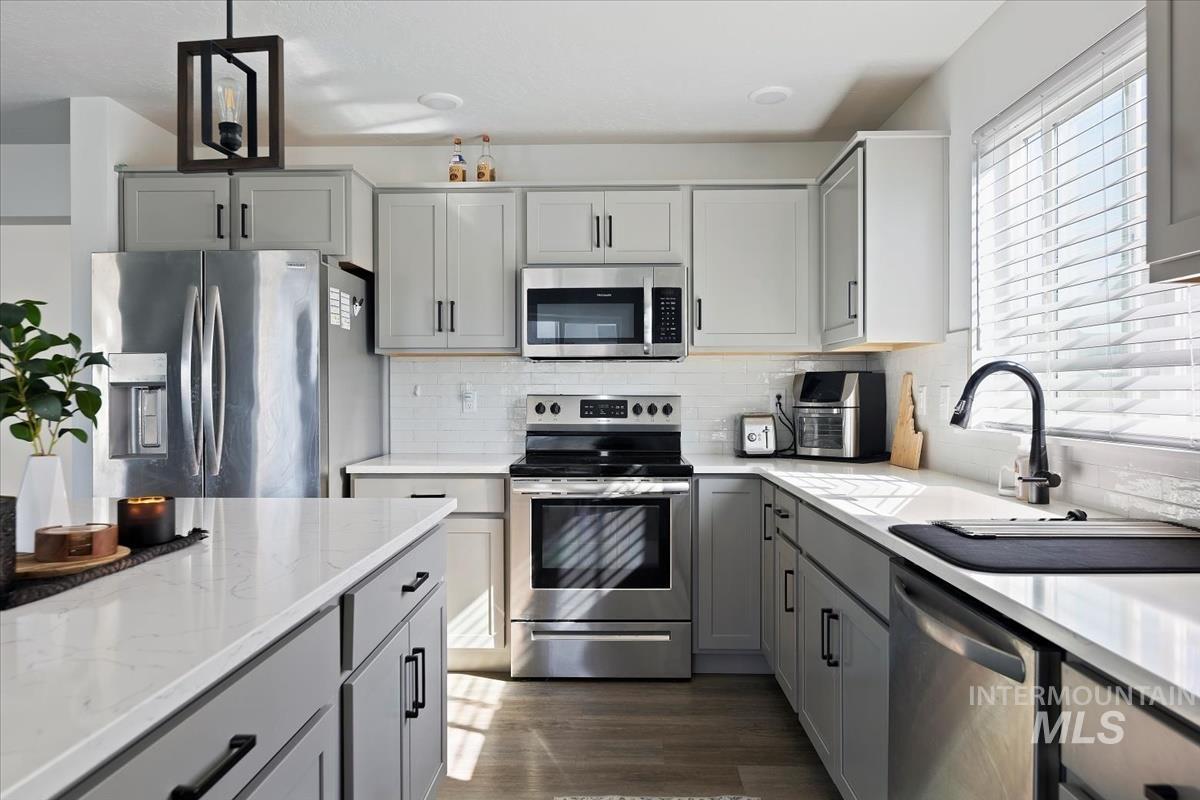Kitchen with gray cabinetry, stainless steel appliances, light stone counters, decorative backsplash, and dark wood-style flooring