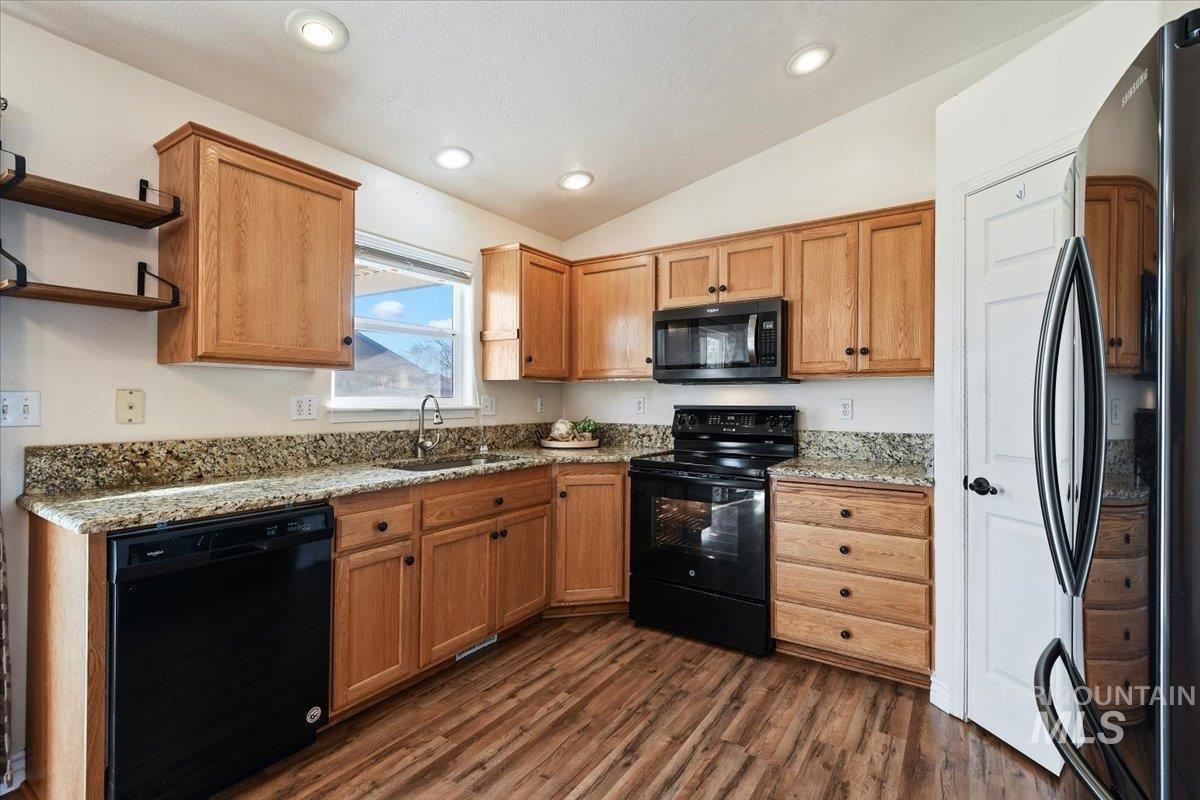 Kitchen with black appliances, light stone counters, lofted ceiling, brown cabinetry, and dark wood finished floors