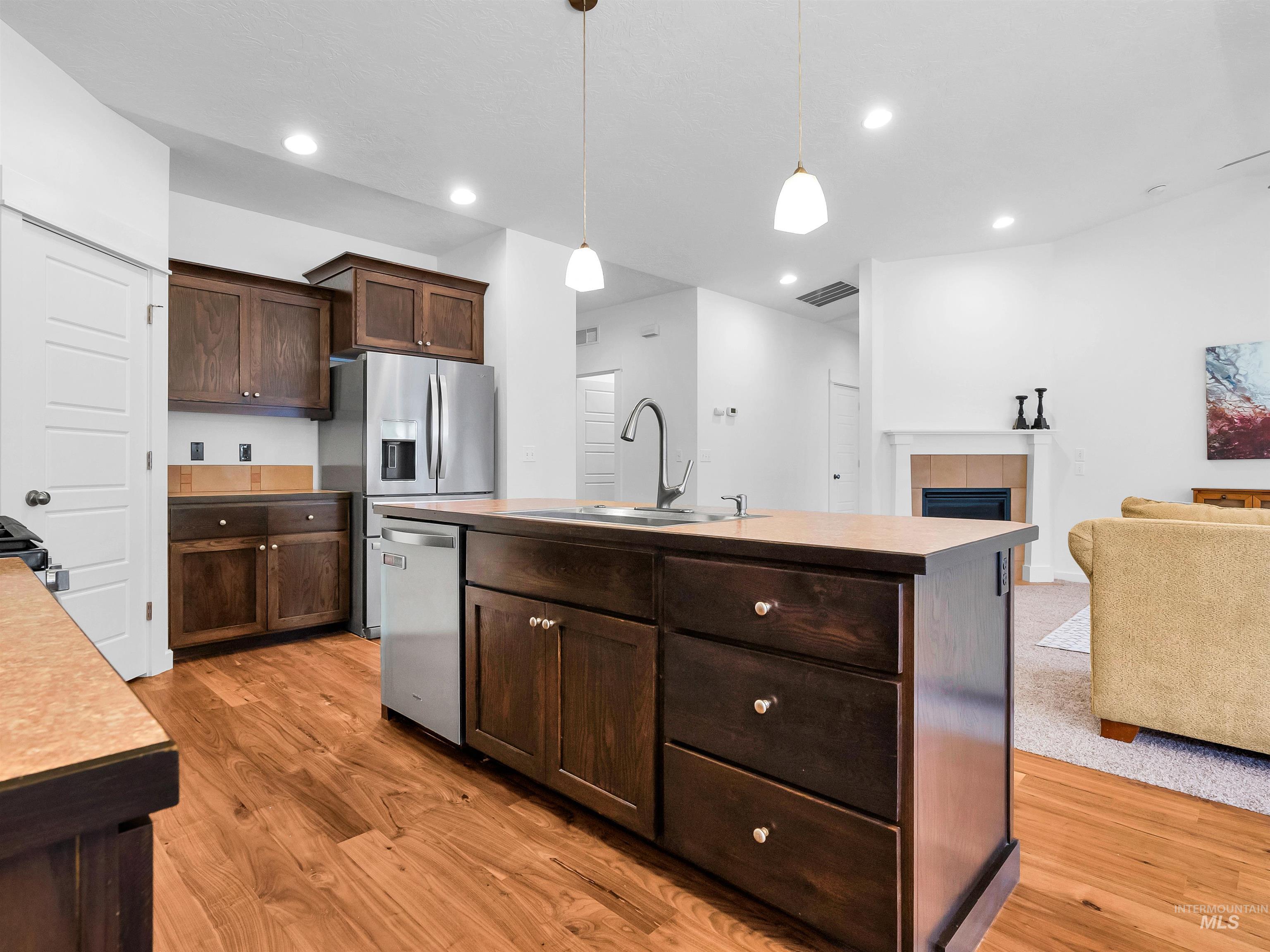 Kitchen featuring dark brown cabinetry, pendant lighting, a tiled fireplace, a center island with sink, and light wood-style floors