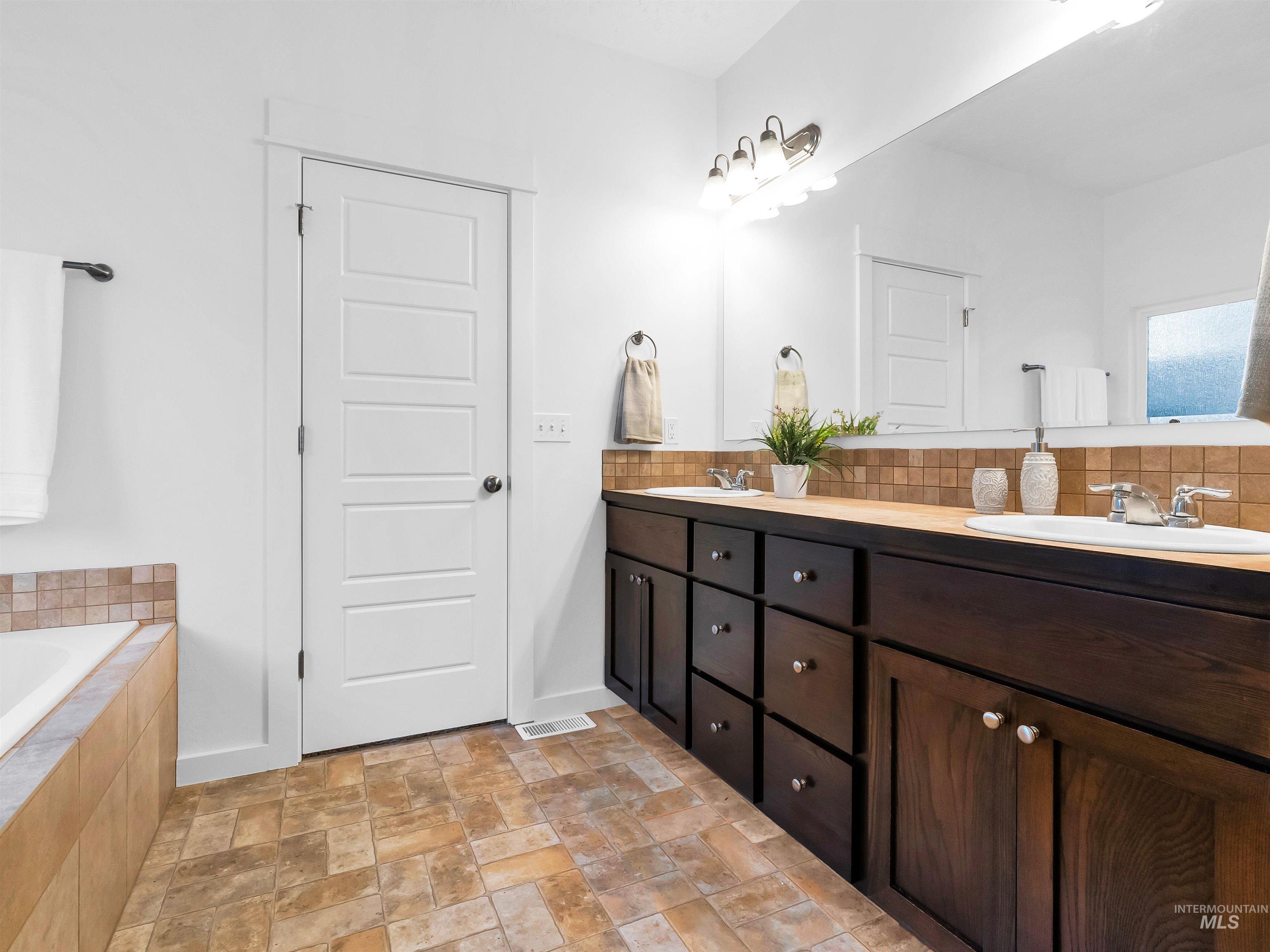 Full bathroom with double vanity, stone finish flooring, decorative backsplash, and tiled tub