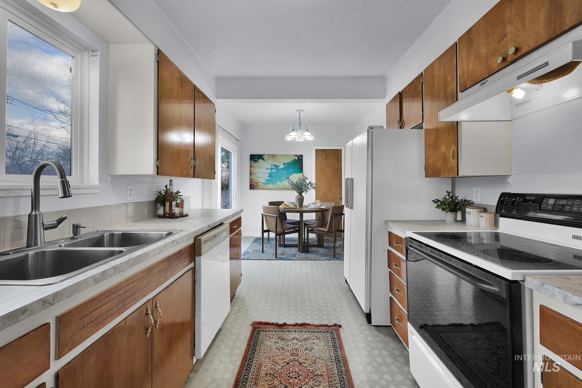 Kitchen with white appliances, light floors, brown cabinetry, under cabinet range hood, and decorative light fixtures