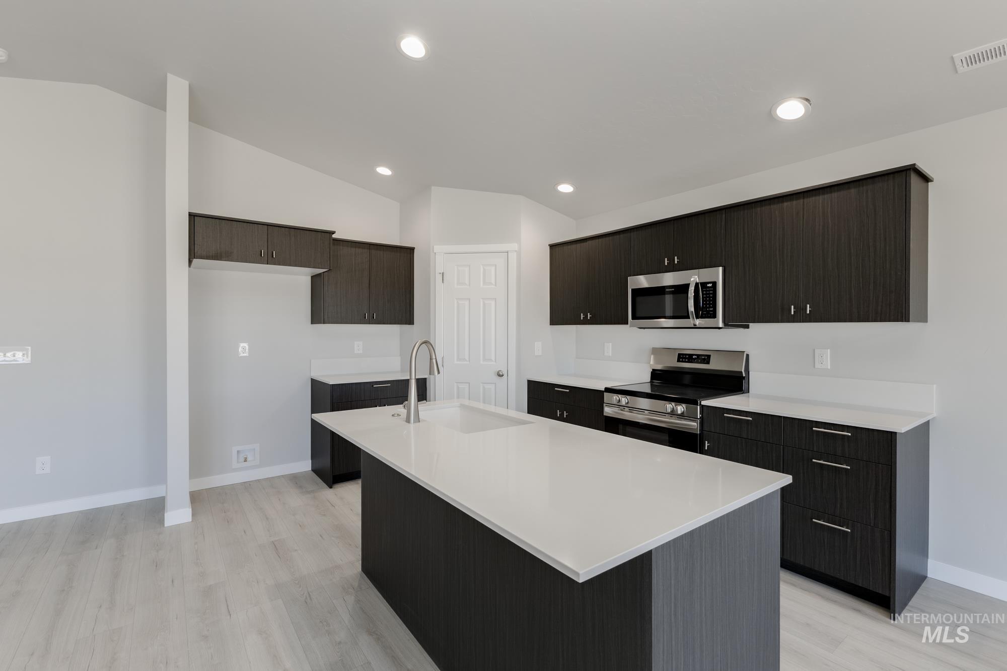 Kitchen with stainless steel appliances, vaulted ceiling, light wood-style floors, a kitchen island with sink, and light stone countertops