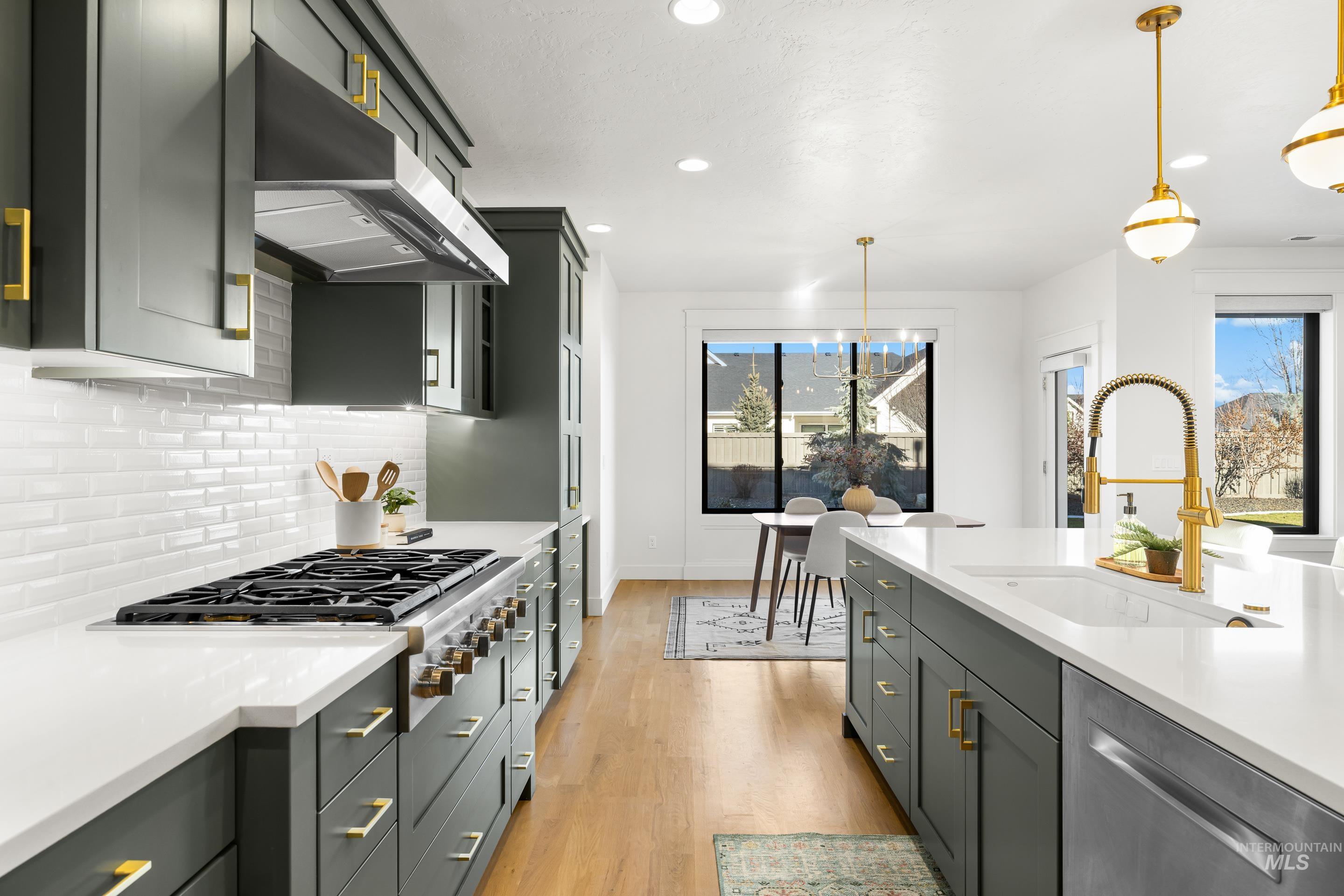 Kitchen with light wood-style flooring, under cabinet range hood, stainless steel appliances, hanging light fixtures, and light stone counters