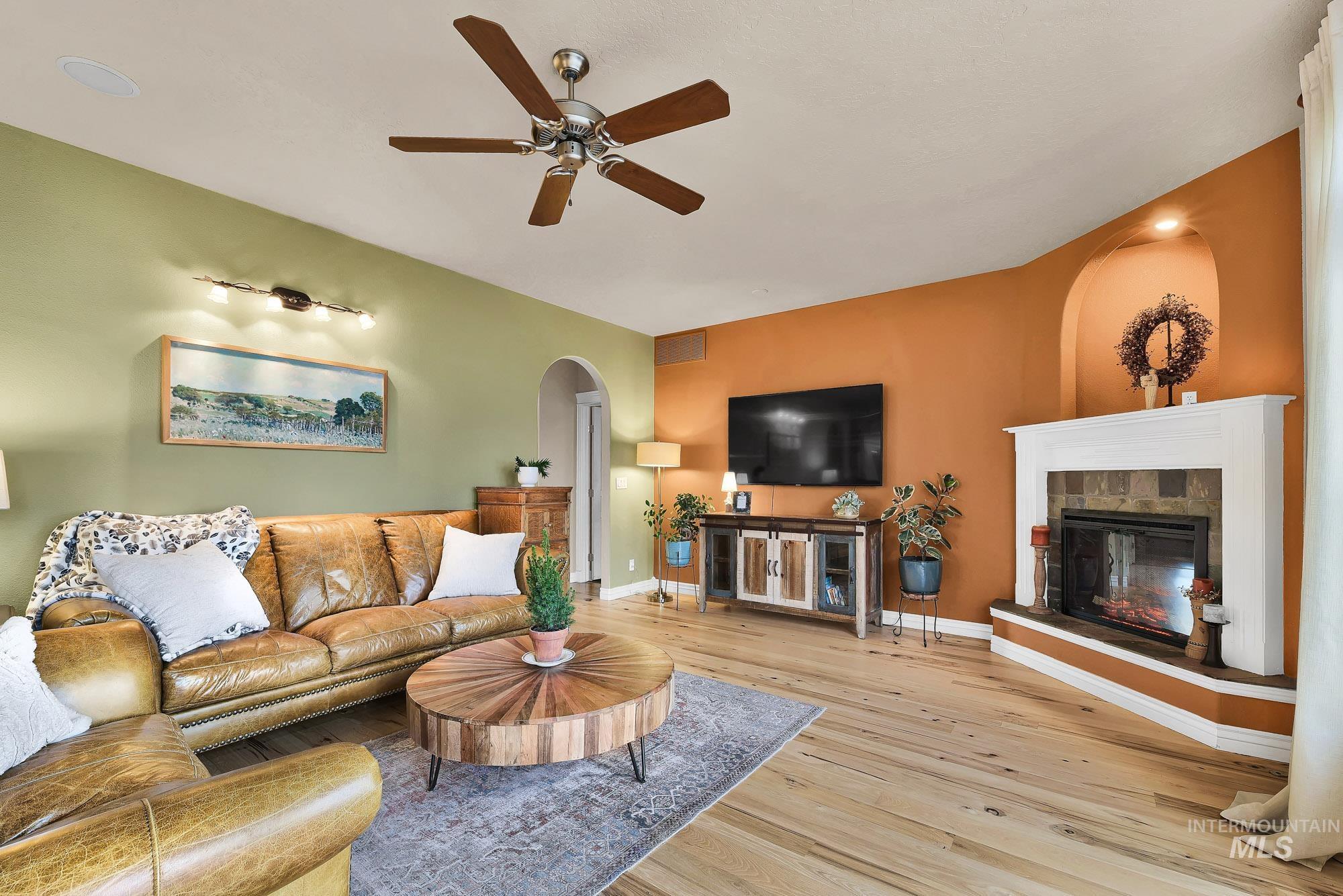 Living area with arched walkways, a tiled fireplace, light wood finished floors, and a ceiling fan