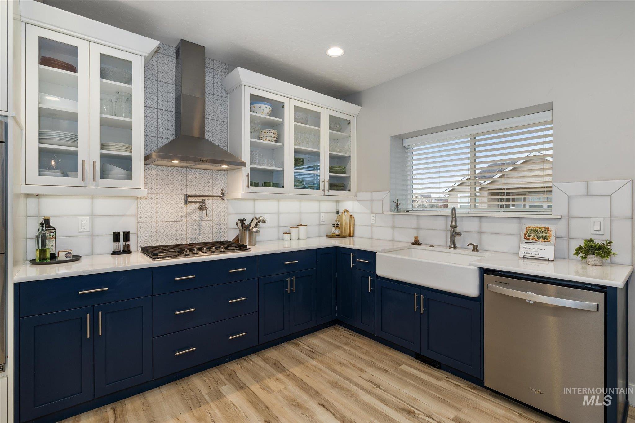 Kitchen featuring wall chimney exhaust hood, blue cabinets, appliances with stainless steel finishes, tasteful backsplash, and light stone counters