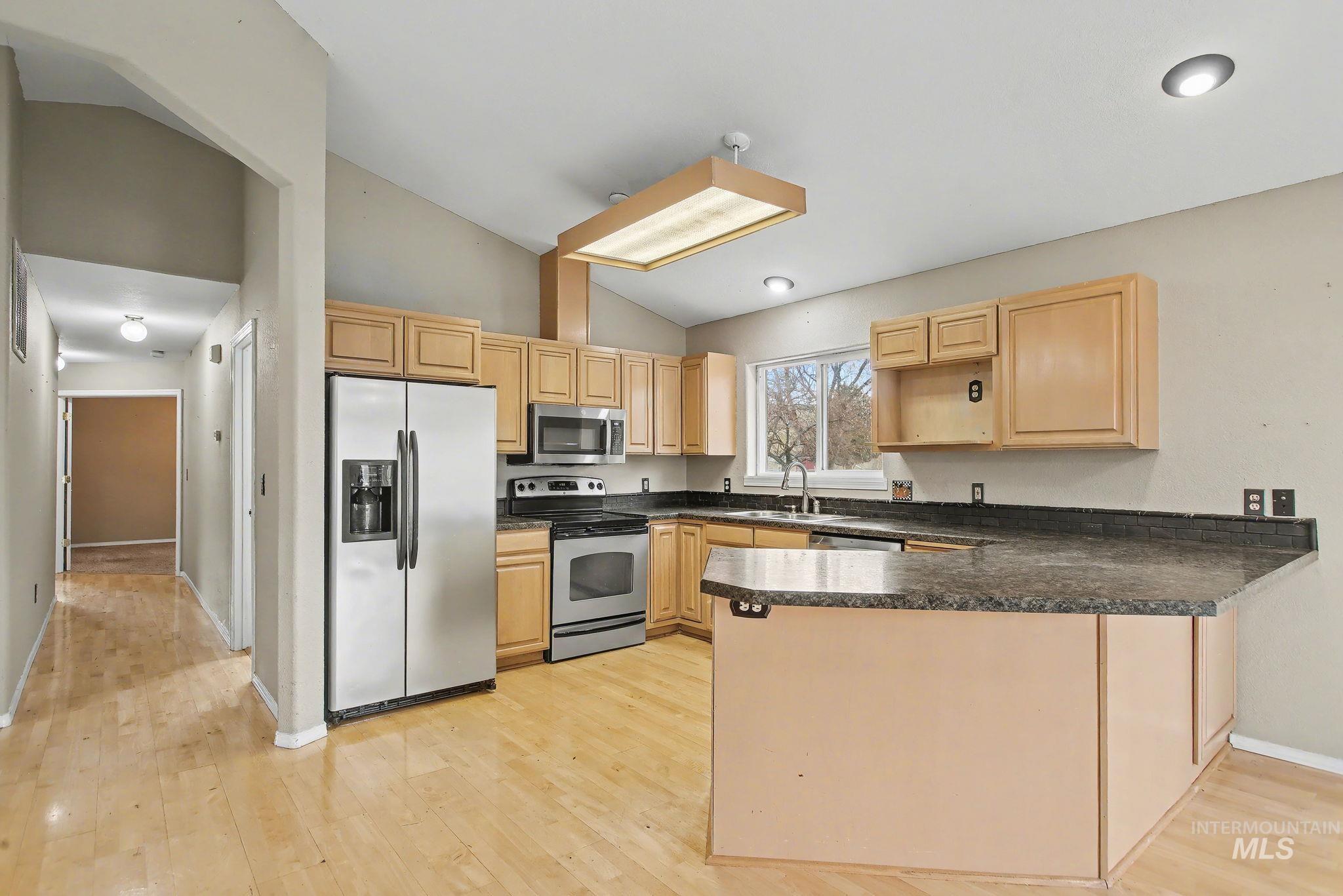 Kitchen with appliances with stainless steel finishes, a peninsula, dark countertops, light brown cabinets, and vaulted ceiling