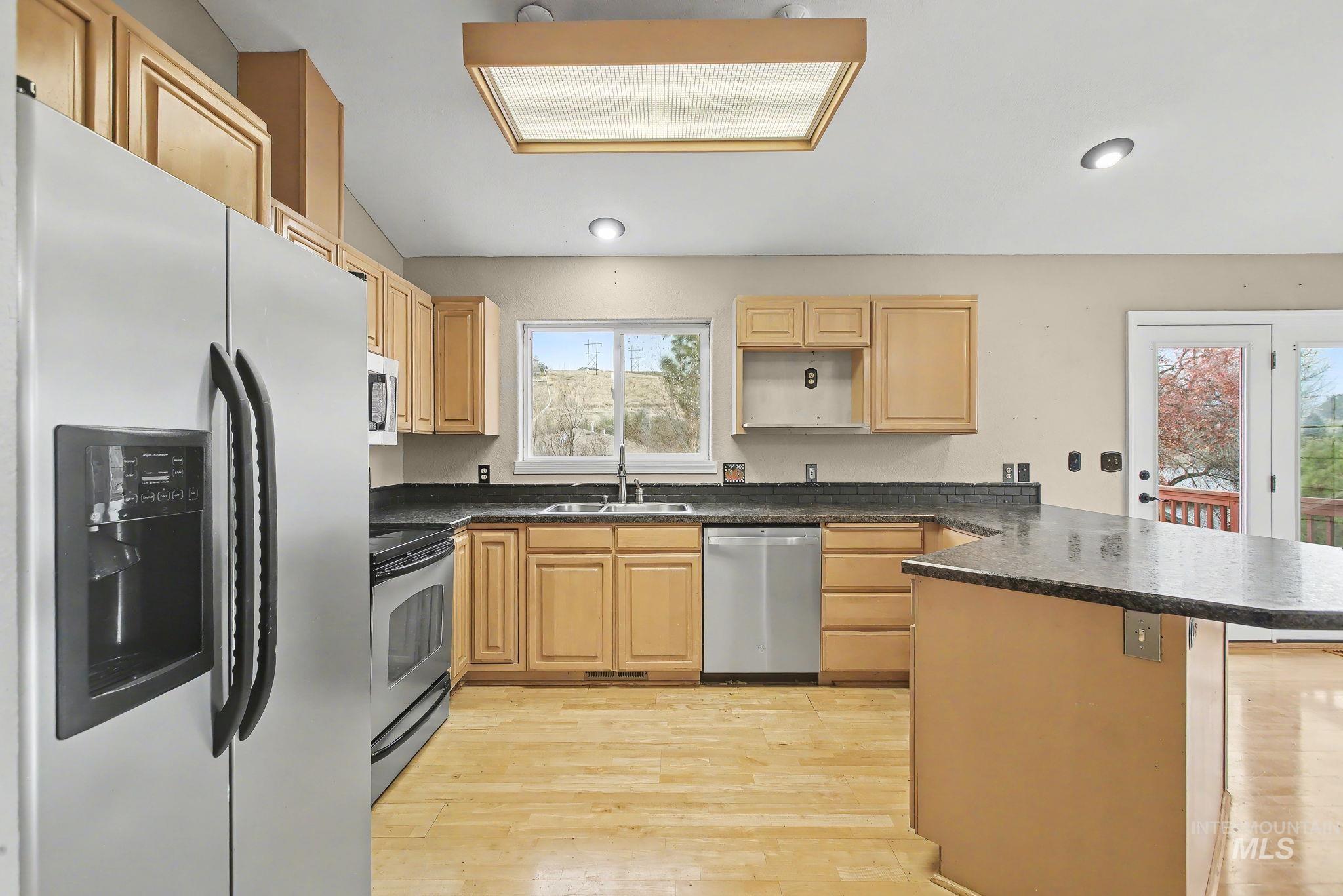 Kitchen with stainless steel appliances, light wood-type flooring, a peninsula, light brown cabinets, and dark stone countertops