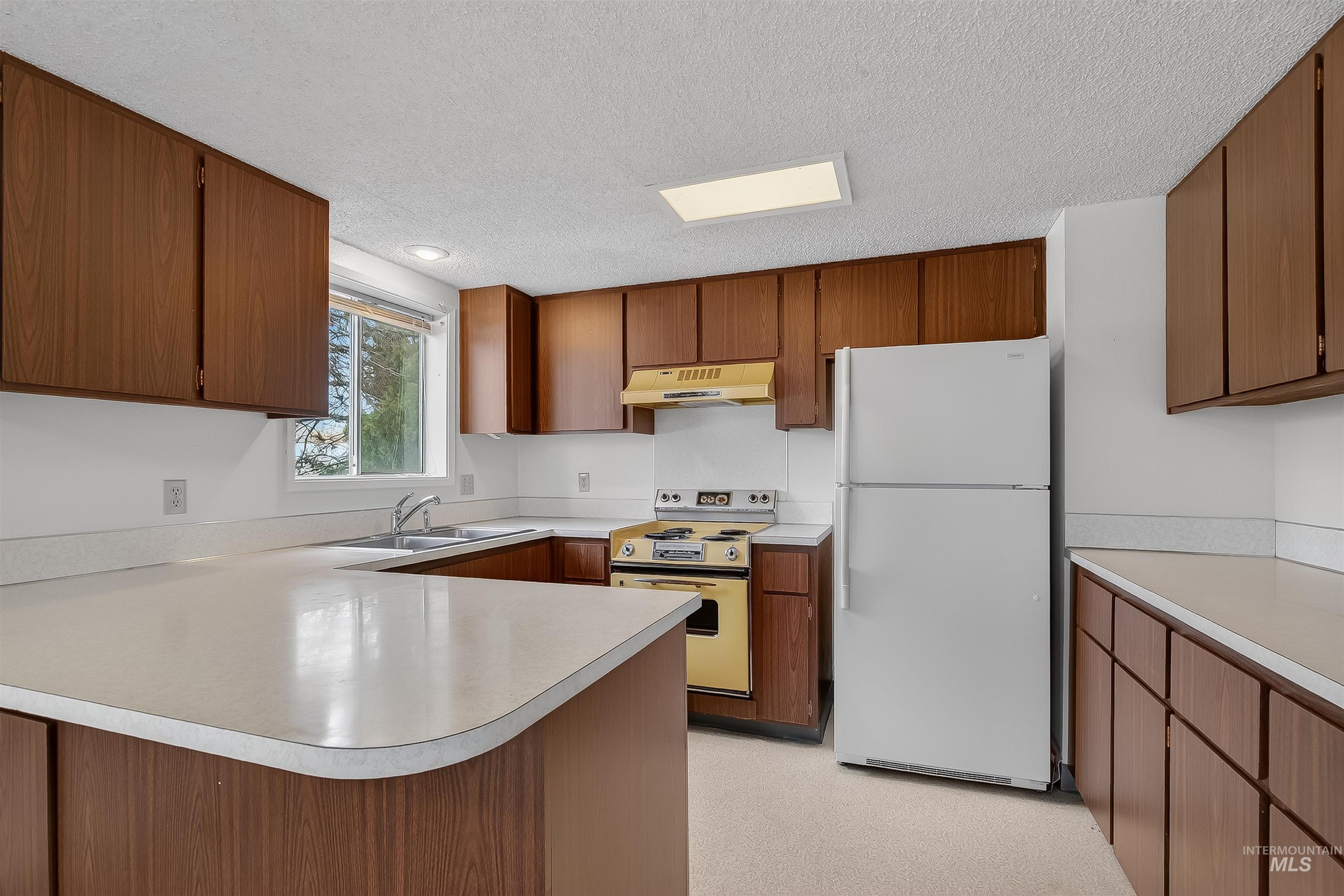 Kitchen featuring freestanding refrigerator, range with electric stovetop, a peninsula, brown cabinets, and a textured ceiling