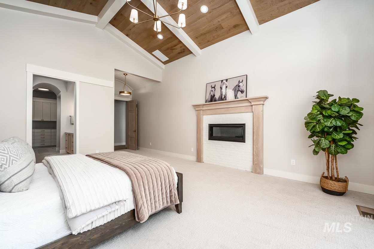 Carpeted bedroom with a glass covered fireplace, ensuite bath, a wooden ceiling with exposed beams, a chandelier, and high vaulted ceiling