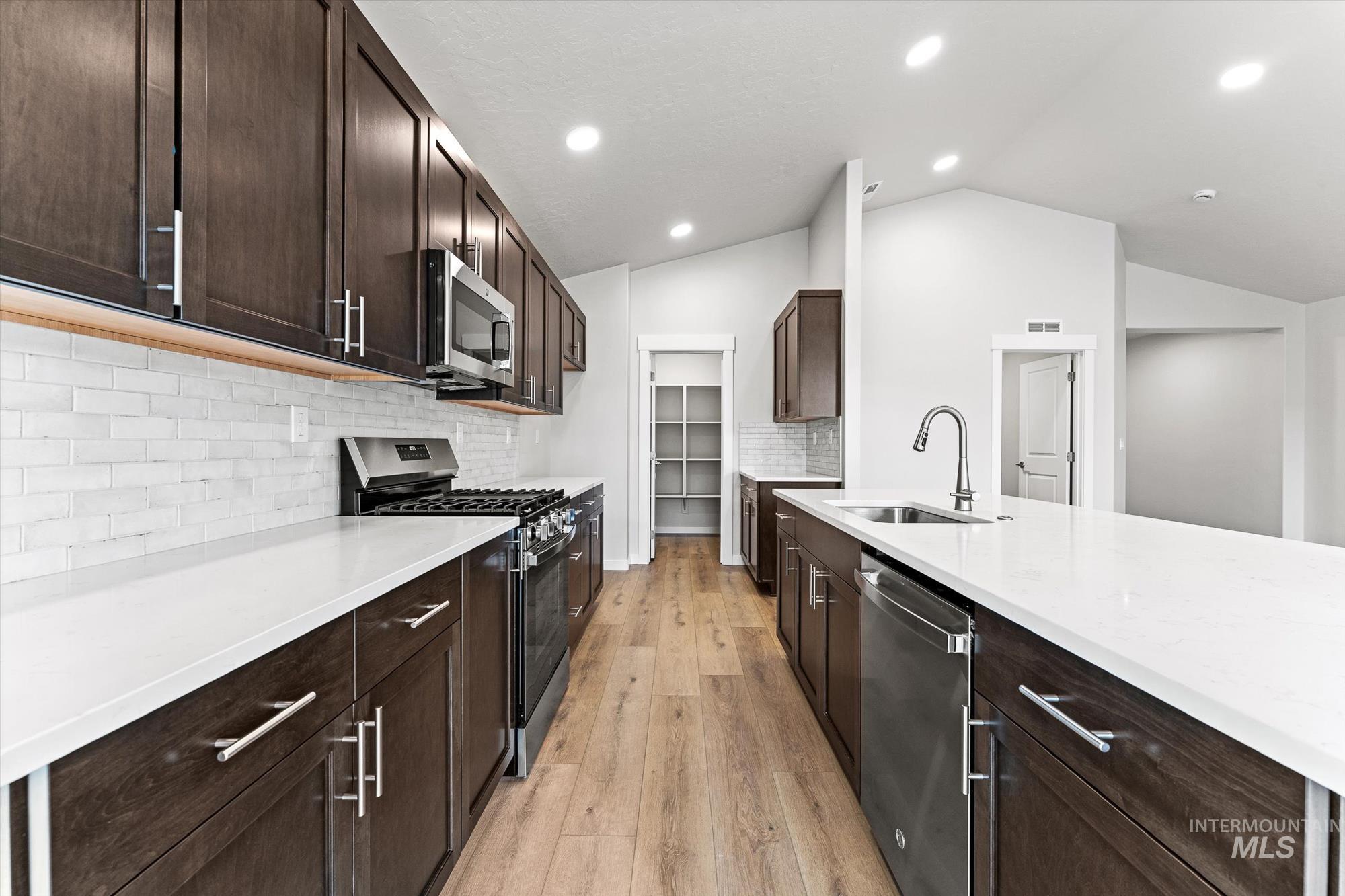 Kitchen featuring dark brown cabinetry, appliances with stainless steel finishes, backsplash, vaulted ceiling, and light stone counters