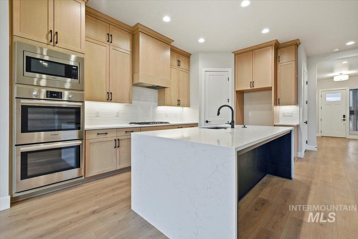 Kitchen featuring light stone counters, appliances with stainless steel finishes, light brown cabinetry, a center island with sink, and light wood-style flooring