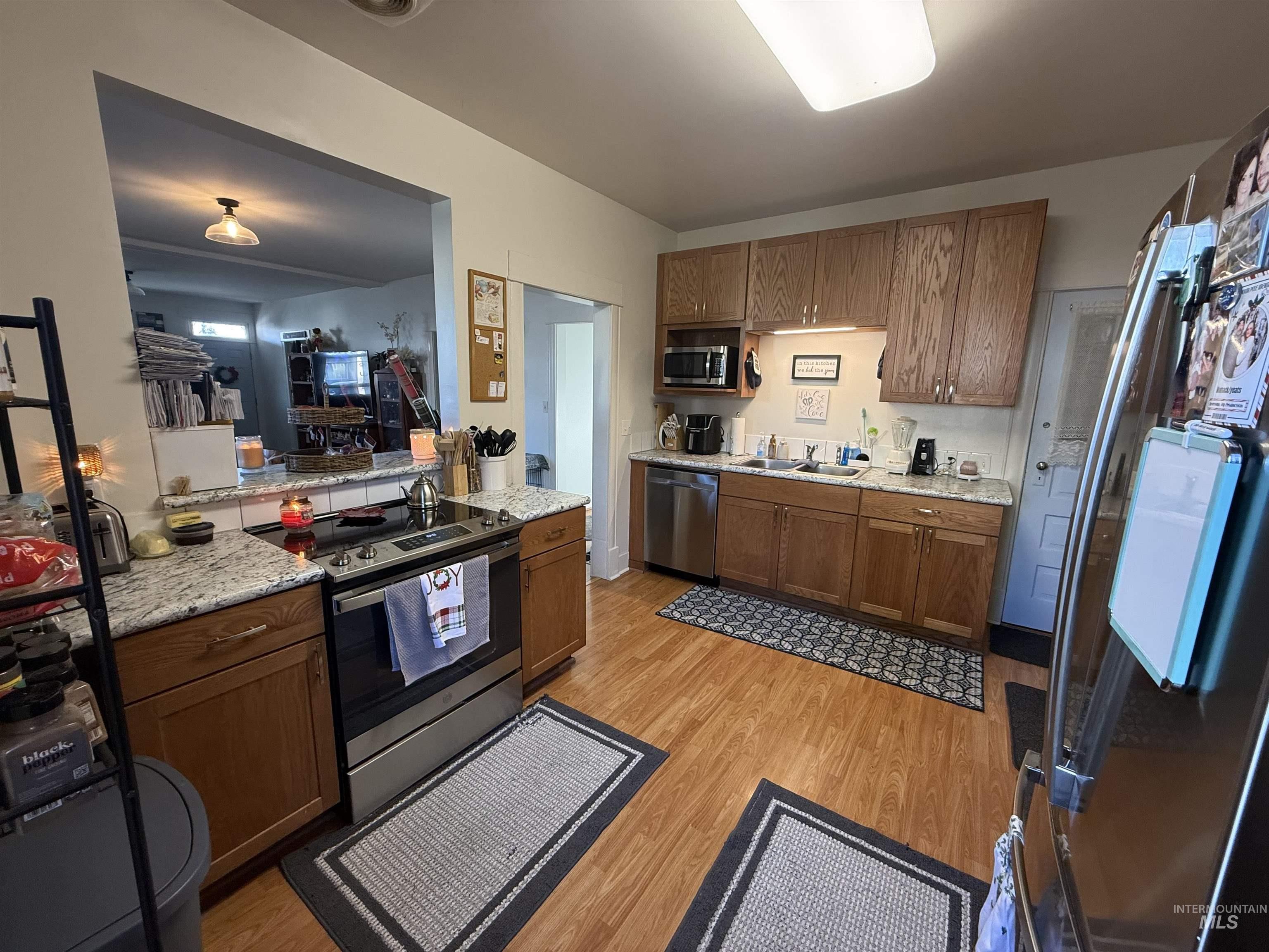Kitchen with stainless steel appliances, brown cabinetry, light wood-style floors, and light stone countertops