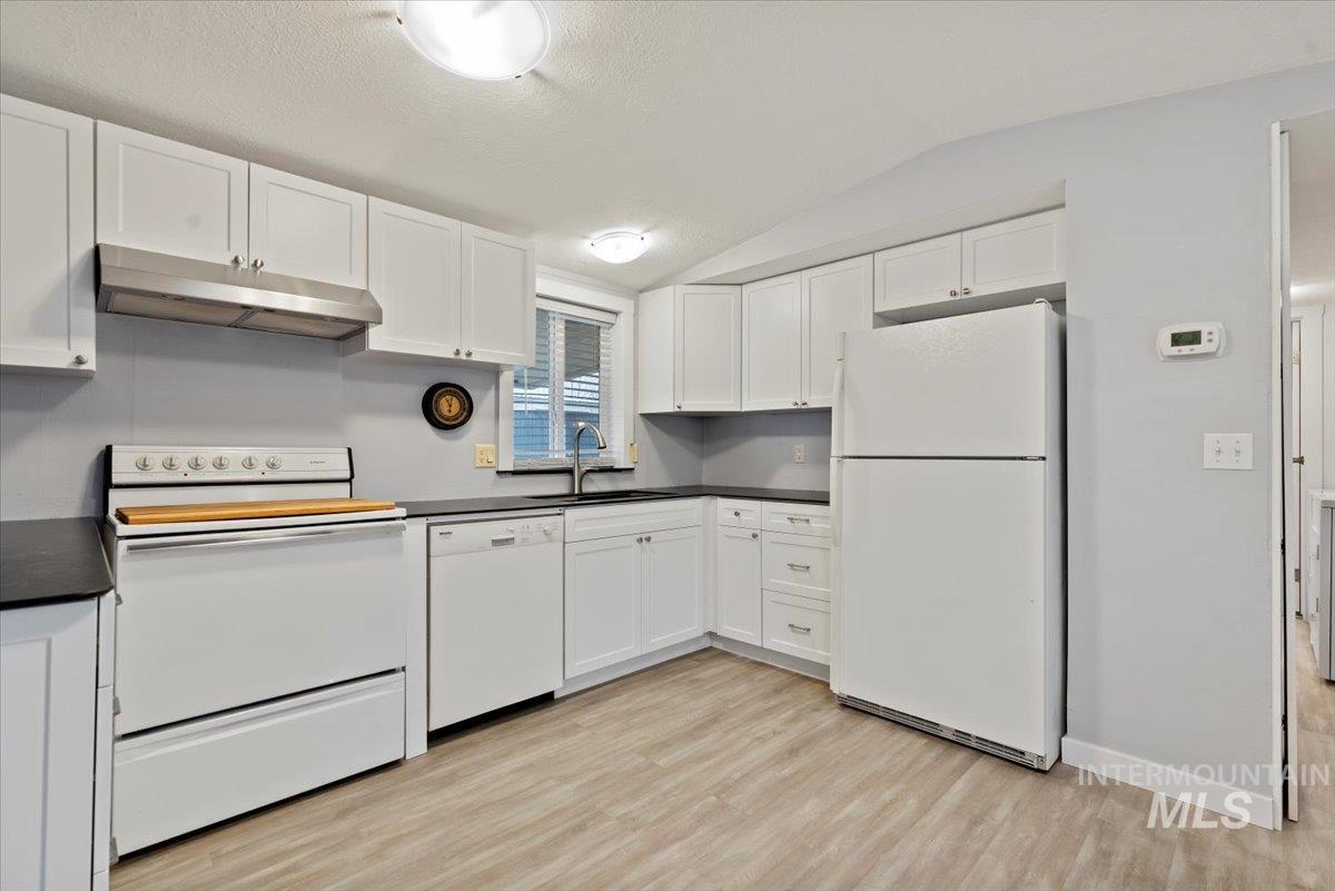 Kitchen featuring white appliances, vaulted ceiling, dark countertops, white cabinets, and under cabinet range hood