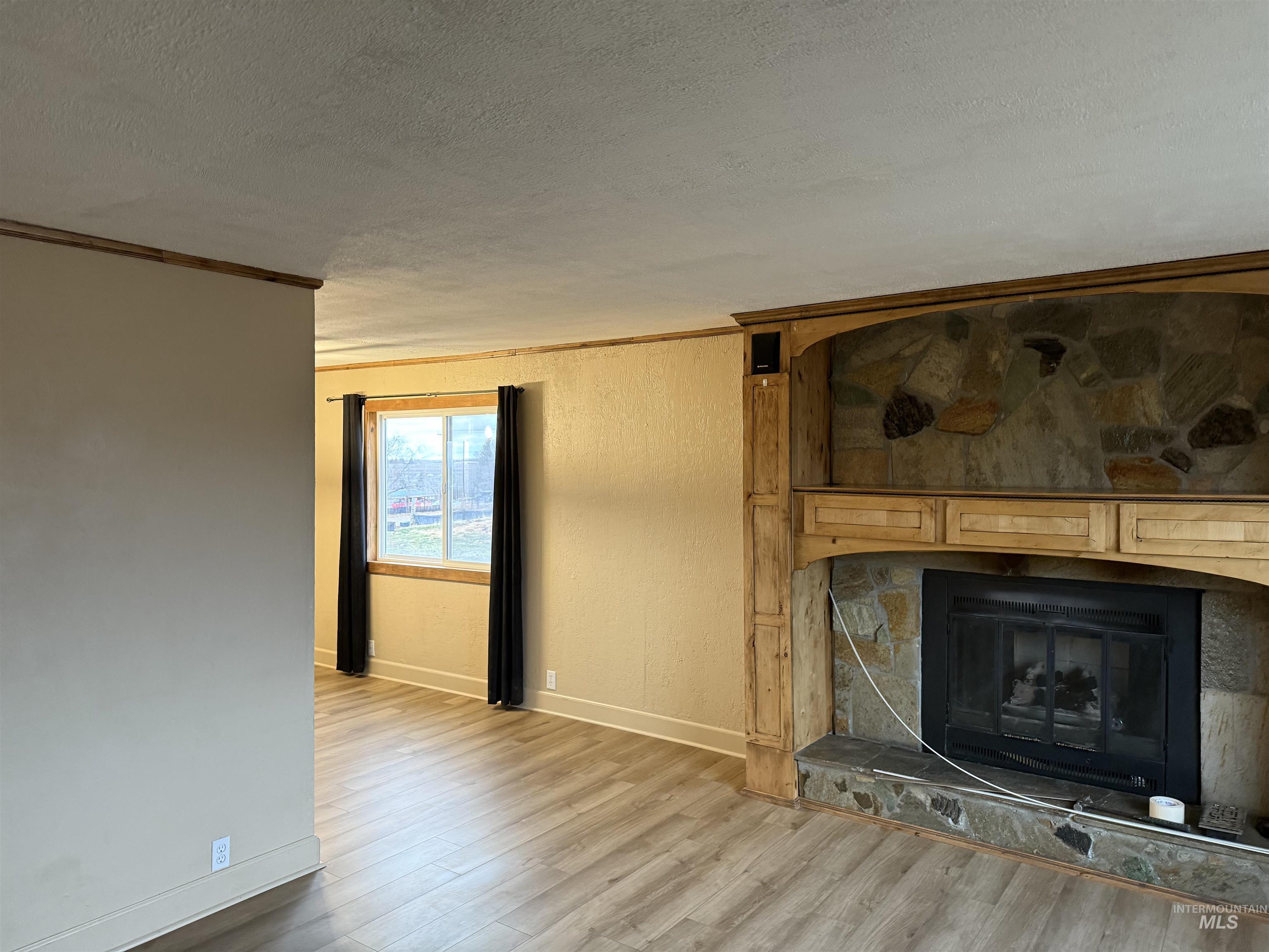 Unfurnished living room with light wood-style floors, crown molding, a stone fireplace, a textured ceiling, and a textured wall