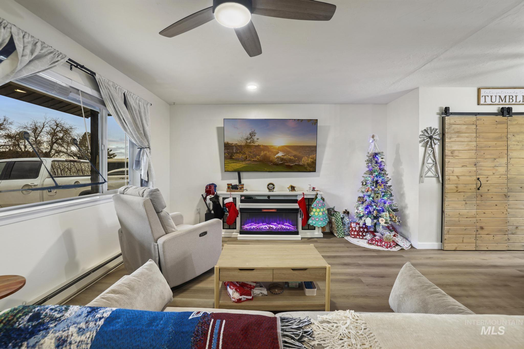 Living room with a baseboard heating unit, wood finished floors, a glass covered fireplace, ceiling fan, and a barn door
