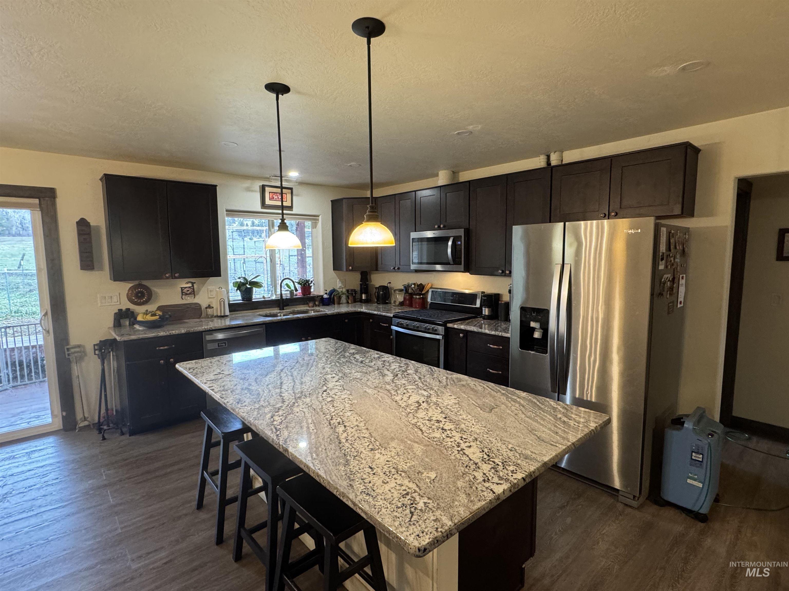 Kitchen with appliances with stainless steel finishes, a kitchen island, a kitchen breakfast bar, light stone countertops, and pendant lighting