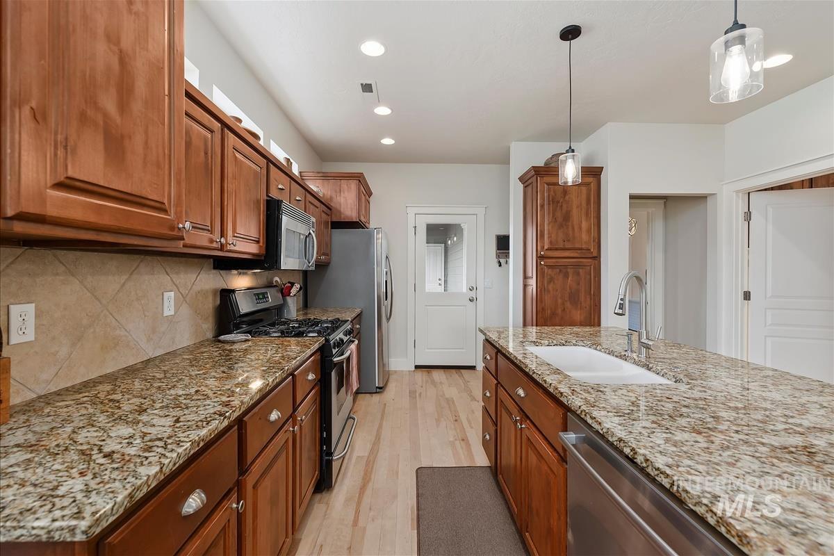 Kitchen with brown cabinets, stainless steel appliances, pendant lighting, light stone counters, and light wood-type flooring