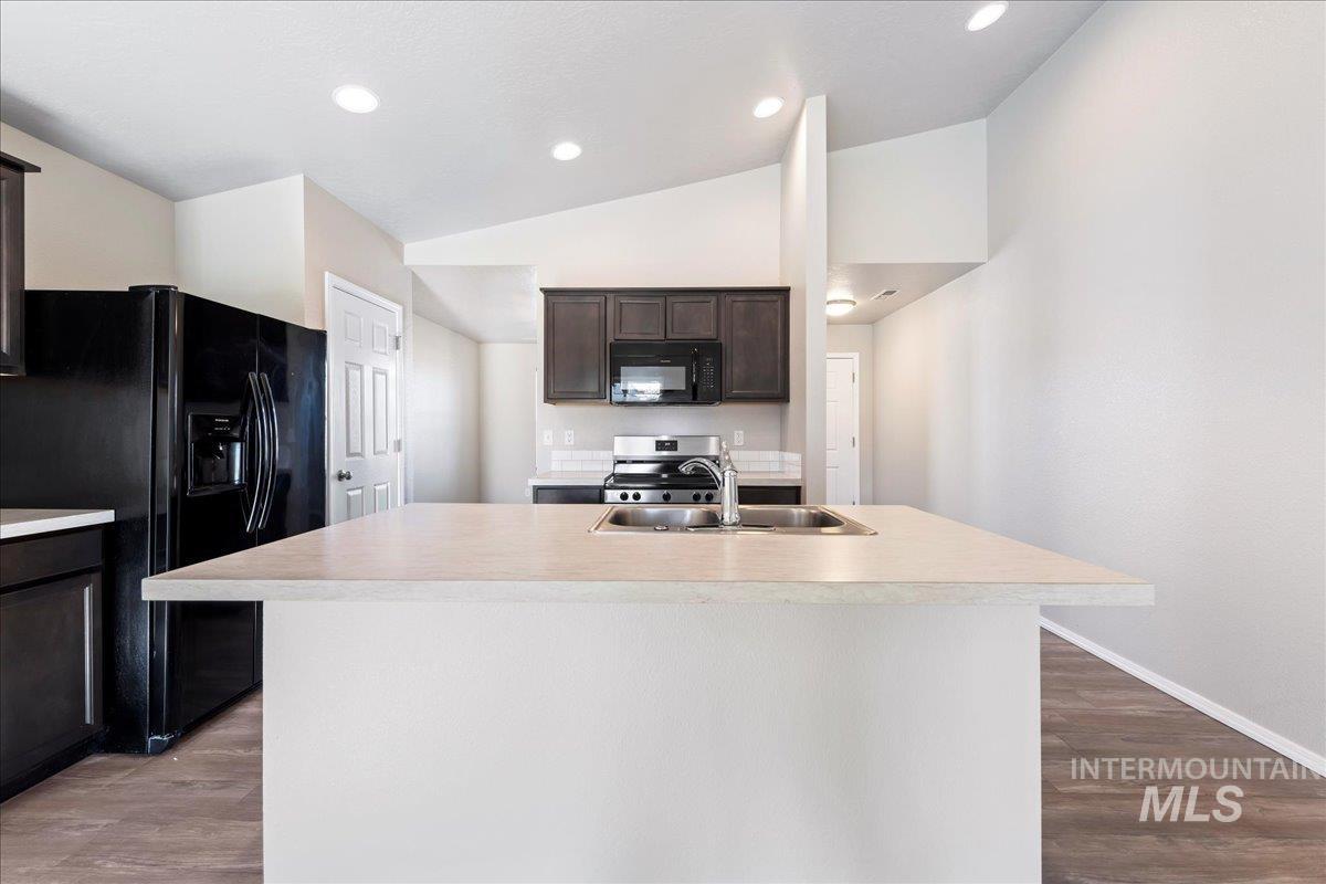 Kitchen with dark brown cabinets, light countertops, black appliances, a kitchen island with sink, and vaulted ceiling