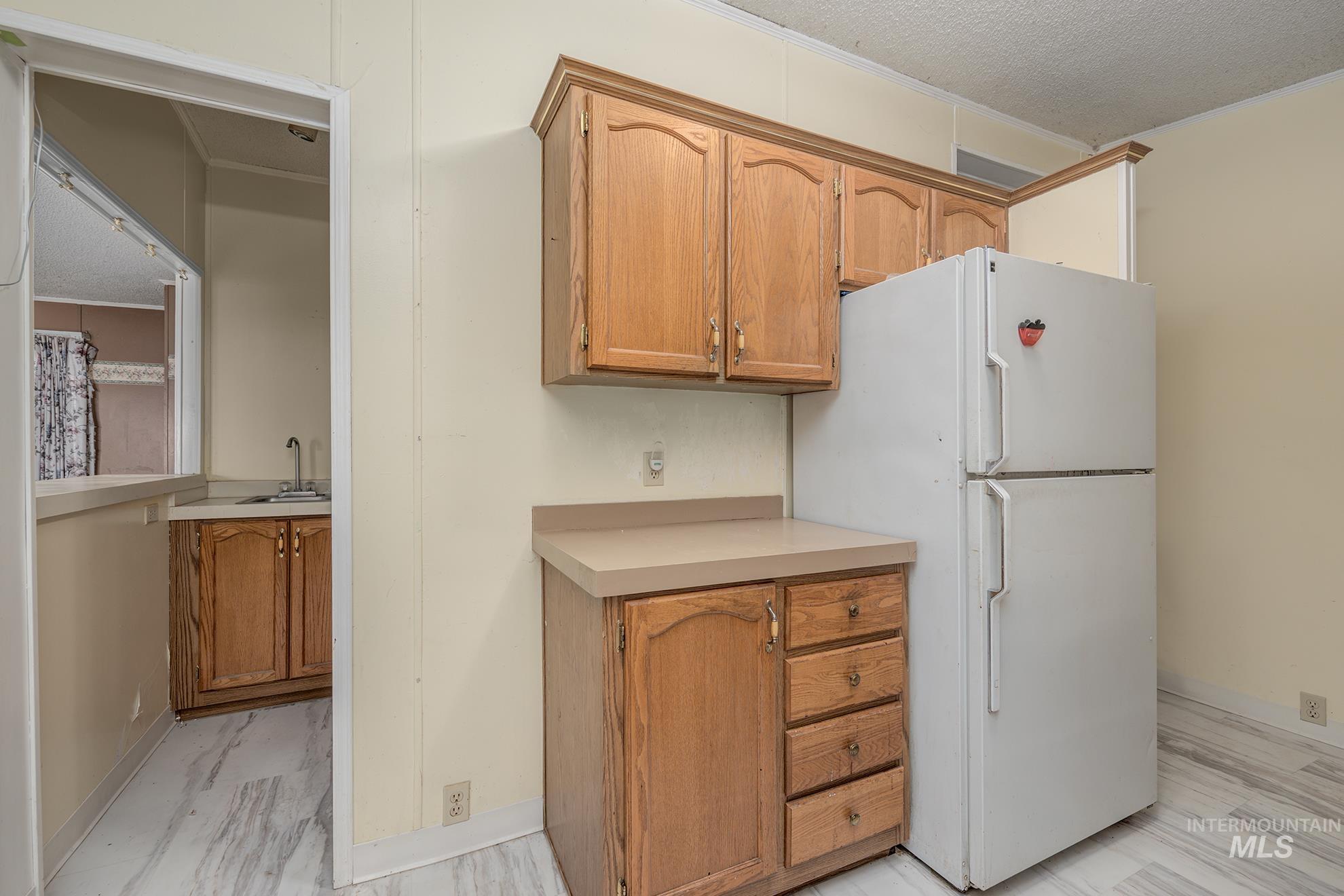 Kitchen with freestanding refrigerator, light countertops, a textured ceiling, brown cabinetry, and light marble finish flooring