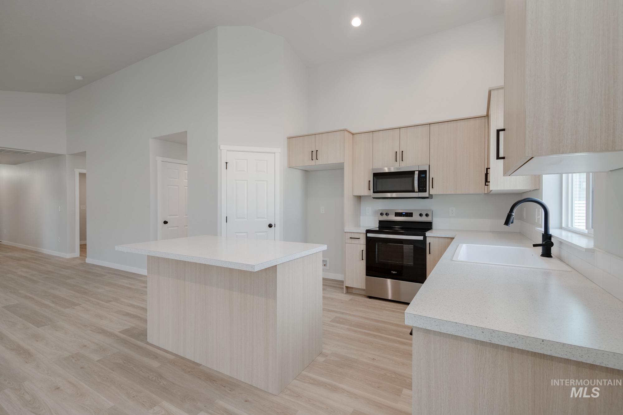 Kitchen featuring light brown cabinets, appliances with stainless steel finishes, a kitchen island, high vaulted ceiling, and light wood finished floors