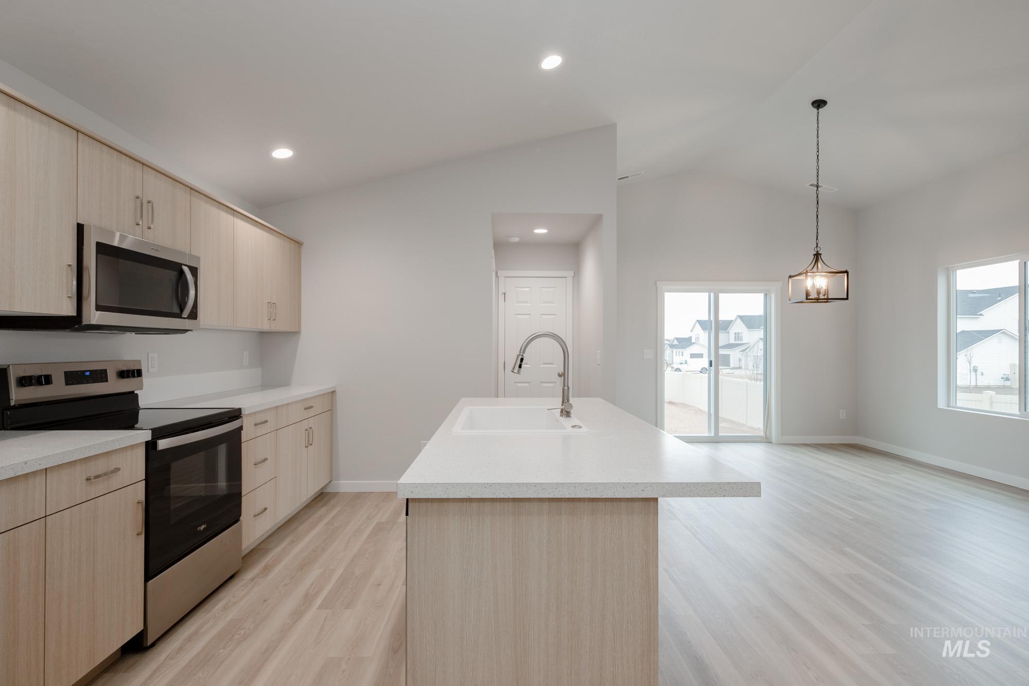 Kitchen with stainless steel appliances, light brown cabinetry, vaulted ceiling, an island with sink, and decorative light fixtures