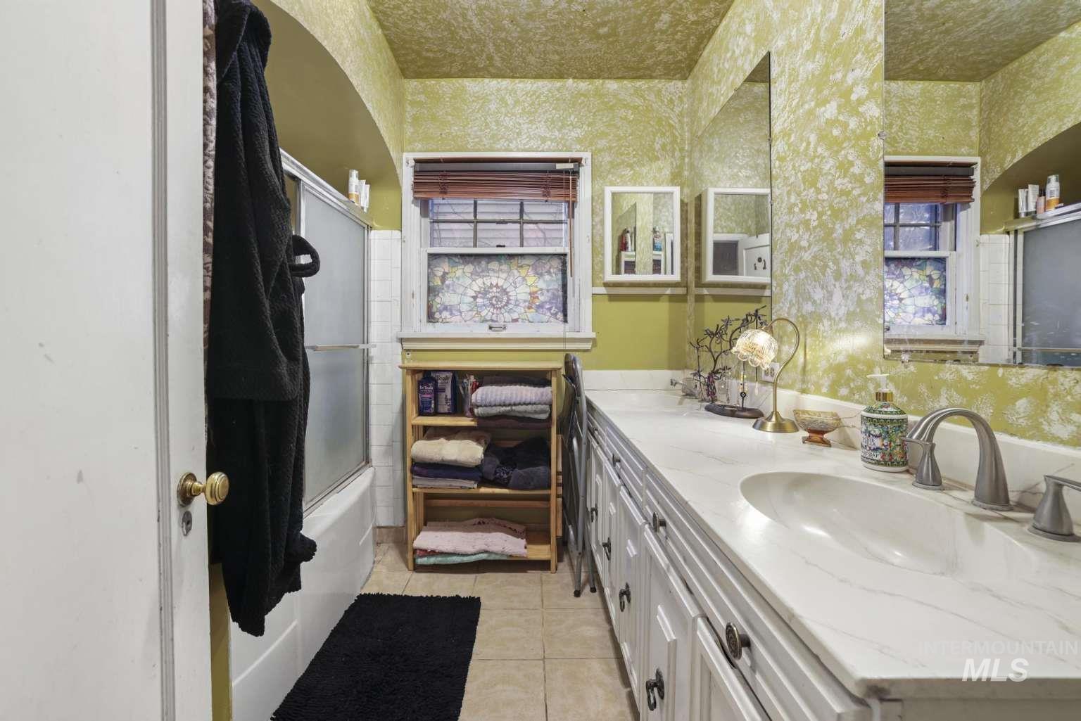 Full bathroom with light tile patterned floors, double vanity, bath / shower combo with glass door, plenty of natural light, and wallpapered walls