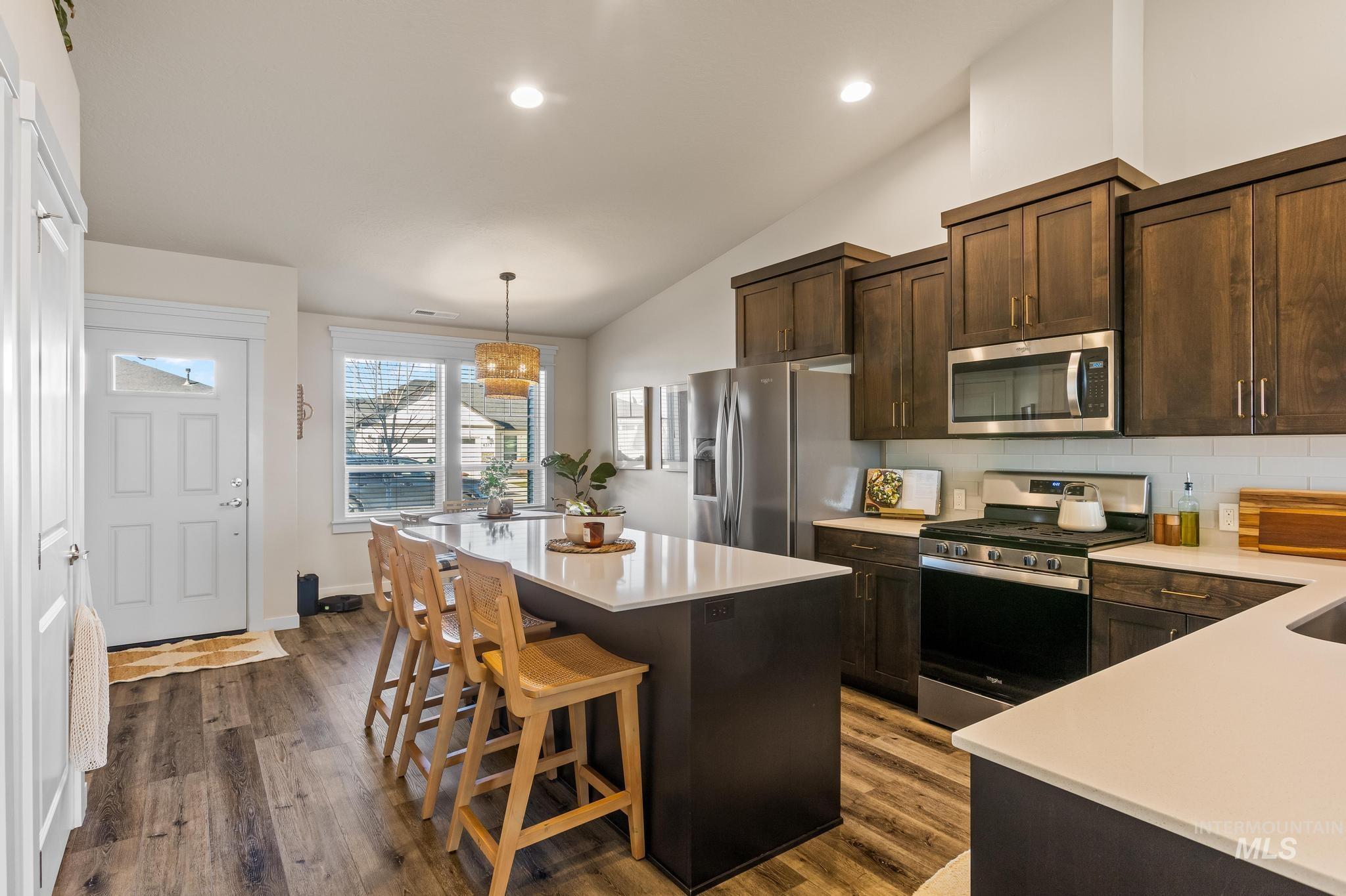 Kitchen with dark brown cabinetry, stainless steel appliances, hanging light fixtures, decorative backsplash, and dark wood-style flooring