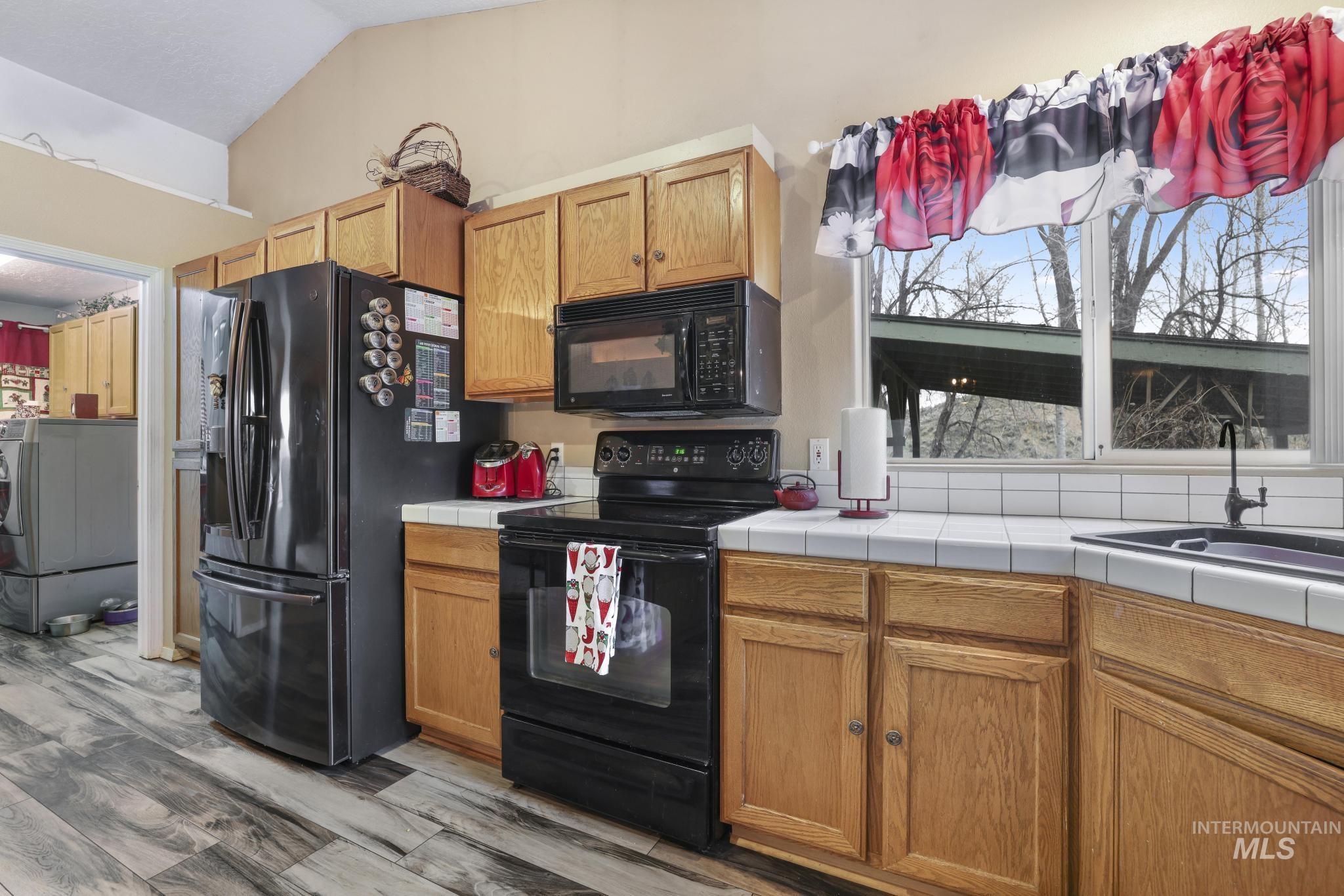 Kitchen with black appliances, tile countertops, light wood finished floors, lofted ceiling, and brown cabinets