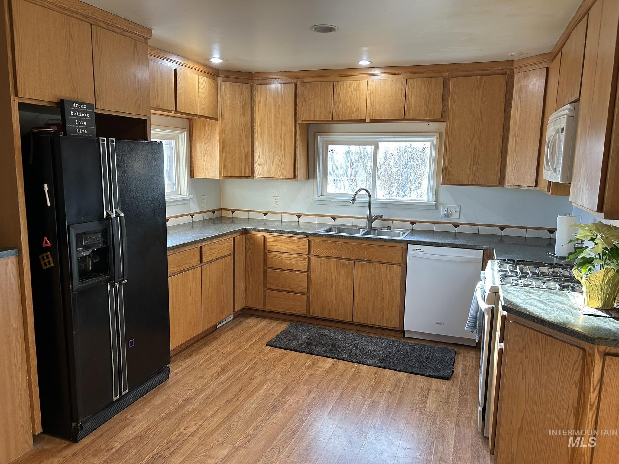 Kitchen with white appliances, brown cabinets, light wood-type flooring, and dark stone counters