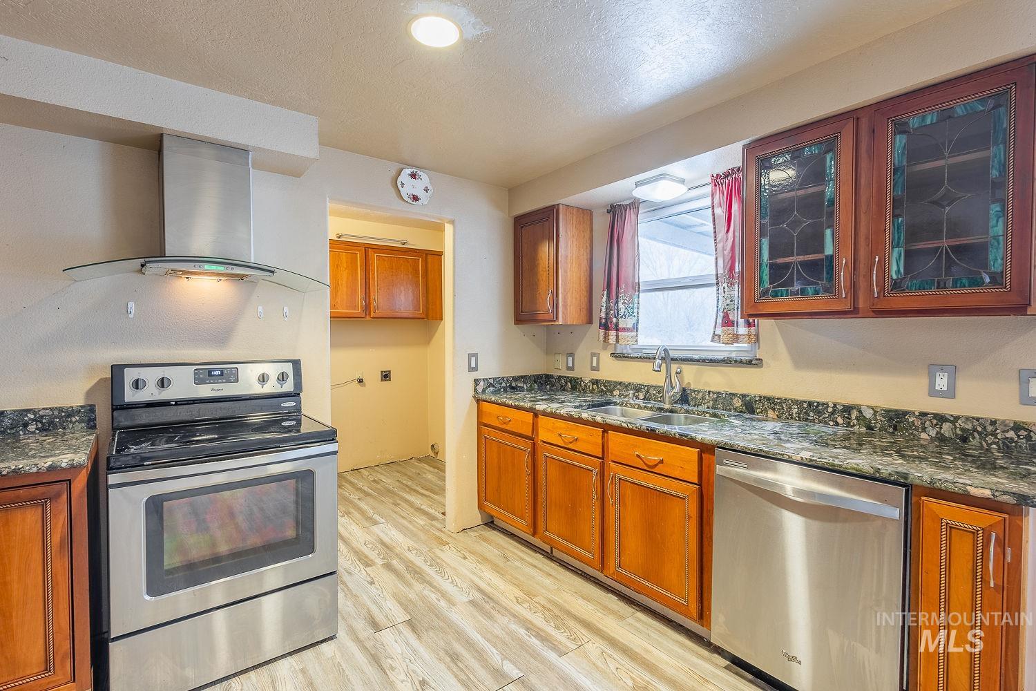 Kitchen with appliances with stainless steel finishes, dark stone countertops, wall chimney exhaust hood, brown cabinetry, and a textured ceiling