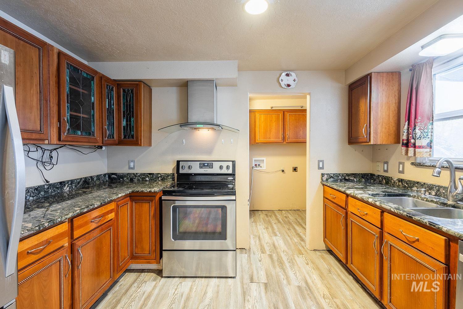 Kitchen featuring stainless steel appliances, dark stone countertops, brown cabinetry, wall chimney exhaust hood, and a textured ceiling