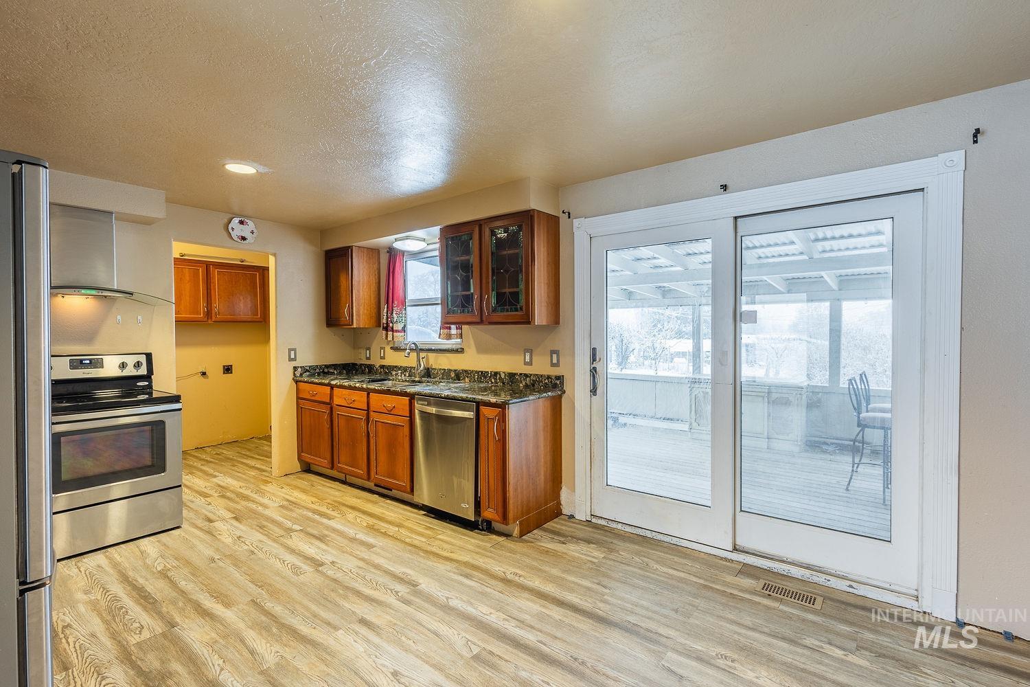 Kitchen featuring stainless steel appliances, brown cabinetry, wall chimney range hood, glass insert cabinets, and light wood finished floors
