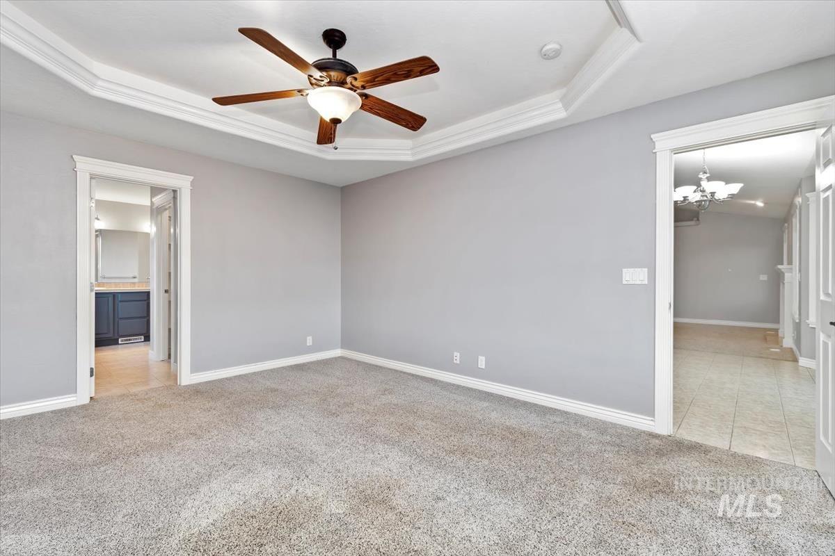 Unfurnished bedroom featuring light carpet, a tray ceiling, a chandelier, ensuite bath, and ceiling fan