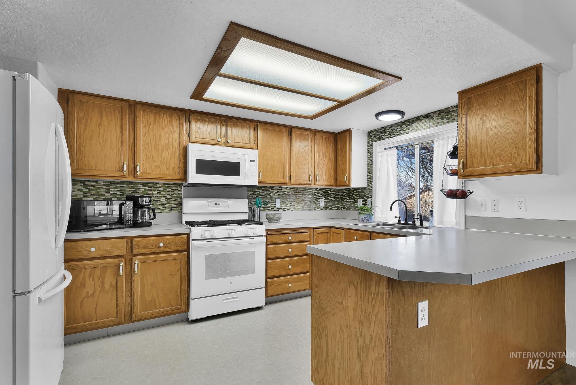 Kitchen featuring white appliances, light floors, brown cabinetry, light countertops, and a peninsula