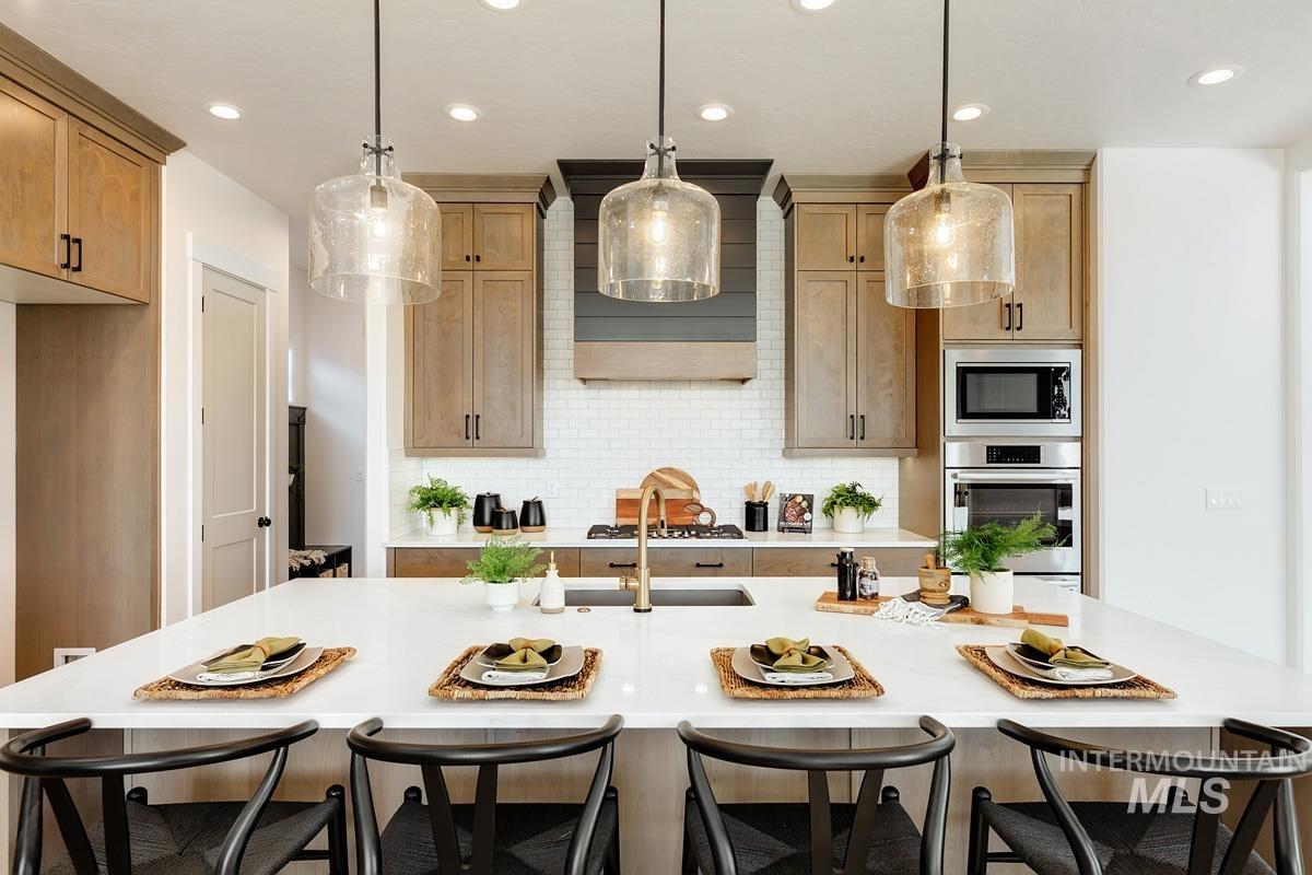 Kitchen with a breakfast bar area, decorative backsplash, appliances with stainless steel finishes, hanging light fixtures, and a kitchen island with sink