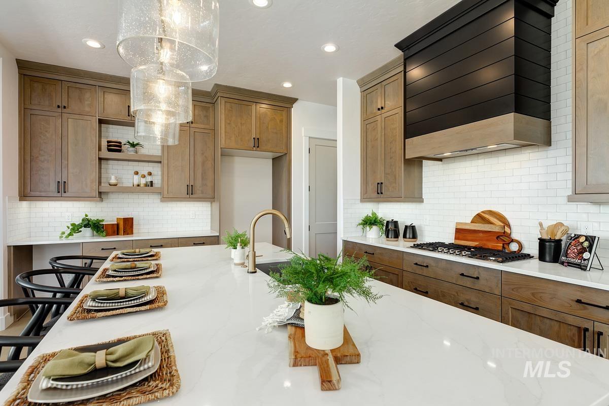 Kitchen with custom range hood, light stone counters, brown cabinetry, backsplash, and hanging light fixtures
