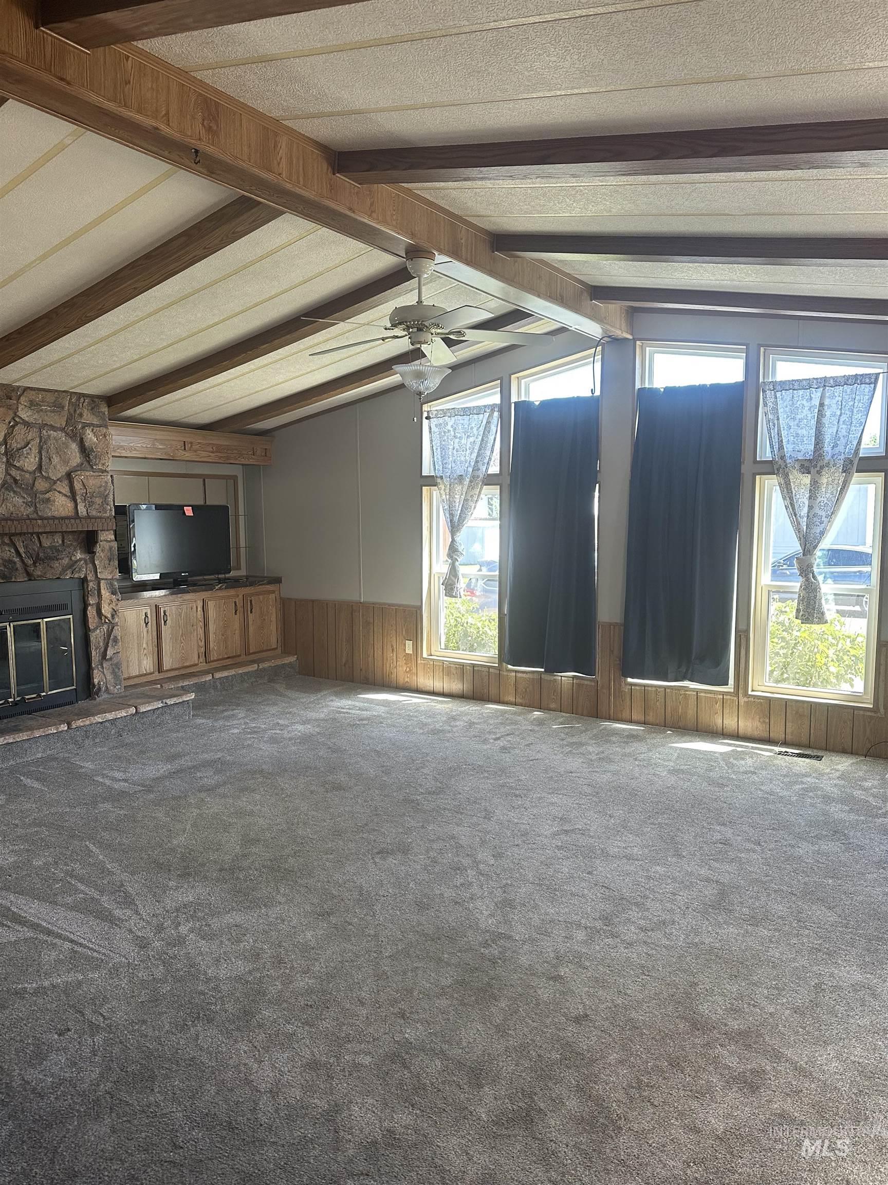 Unfurnished living room featuring carpet flooring, ceiling fan, a stone fireplace, wood walls, and wainscoting