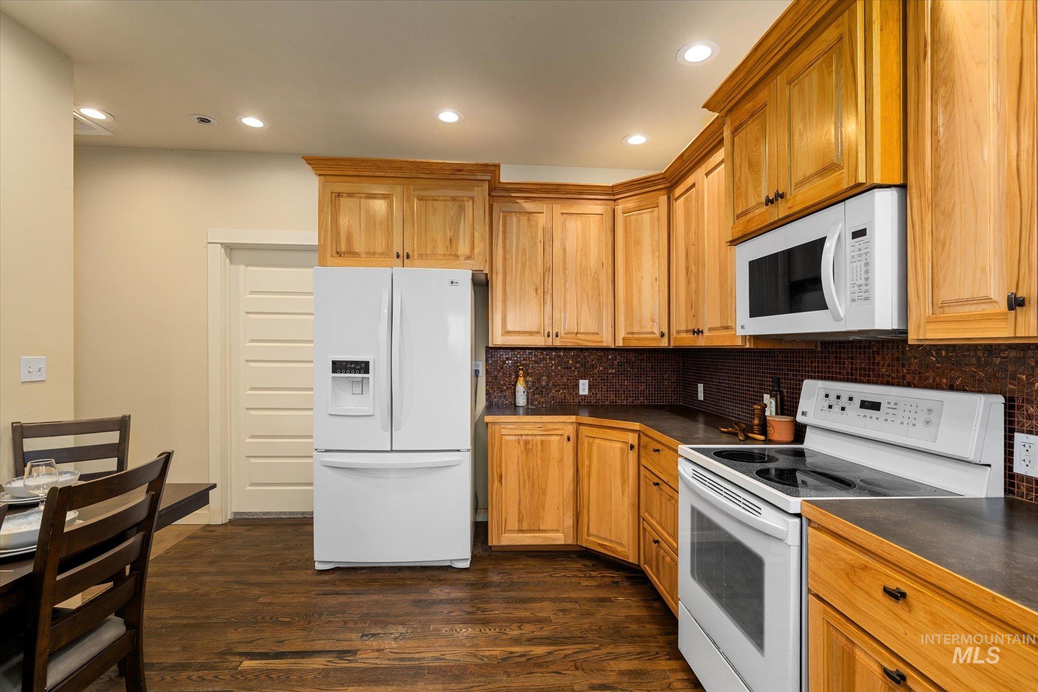 Kitchen with white appliances, dark wood-style flooring, dark countertops, recessed lighting, and tasteful backsplash