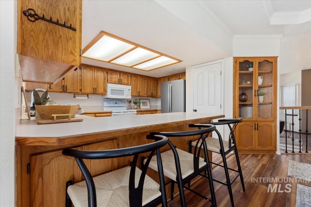 Kitchen featuring light countertops, brown cabinetry, dark wood-style floors, white appliances, and crown molding