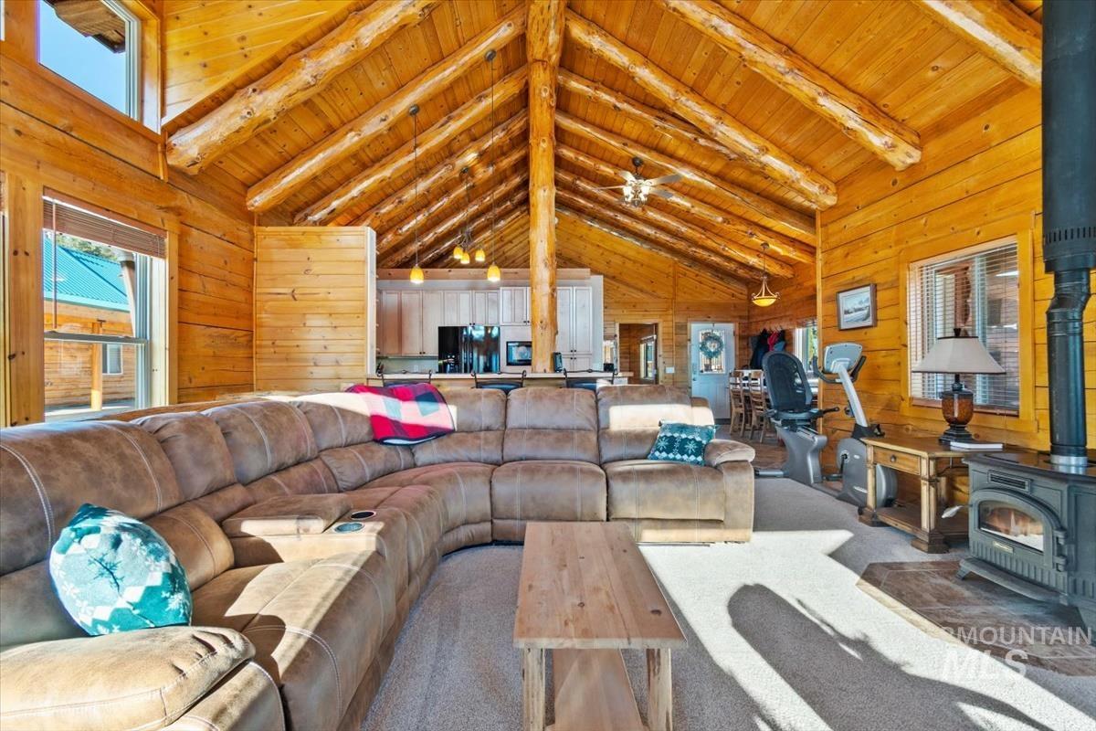 Carpeted living area with wooden walls, a wood stove, a wood ceiling with exposed beams, high vaulted ceiling, and a ceiling fan
