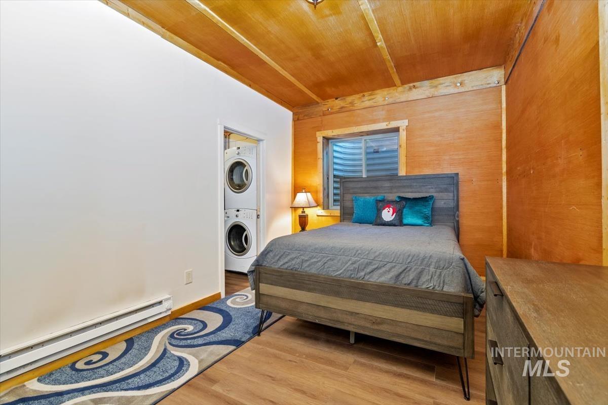 Bedroom with wooden walls, light wood-style floors, baseboard heating, stacked washing machine and dryer, and wooden ceiling