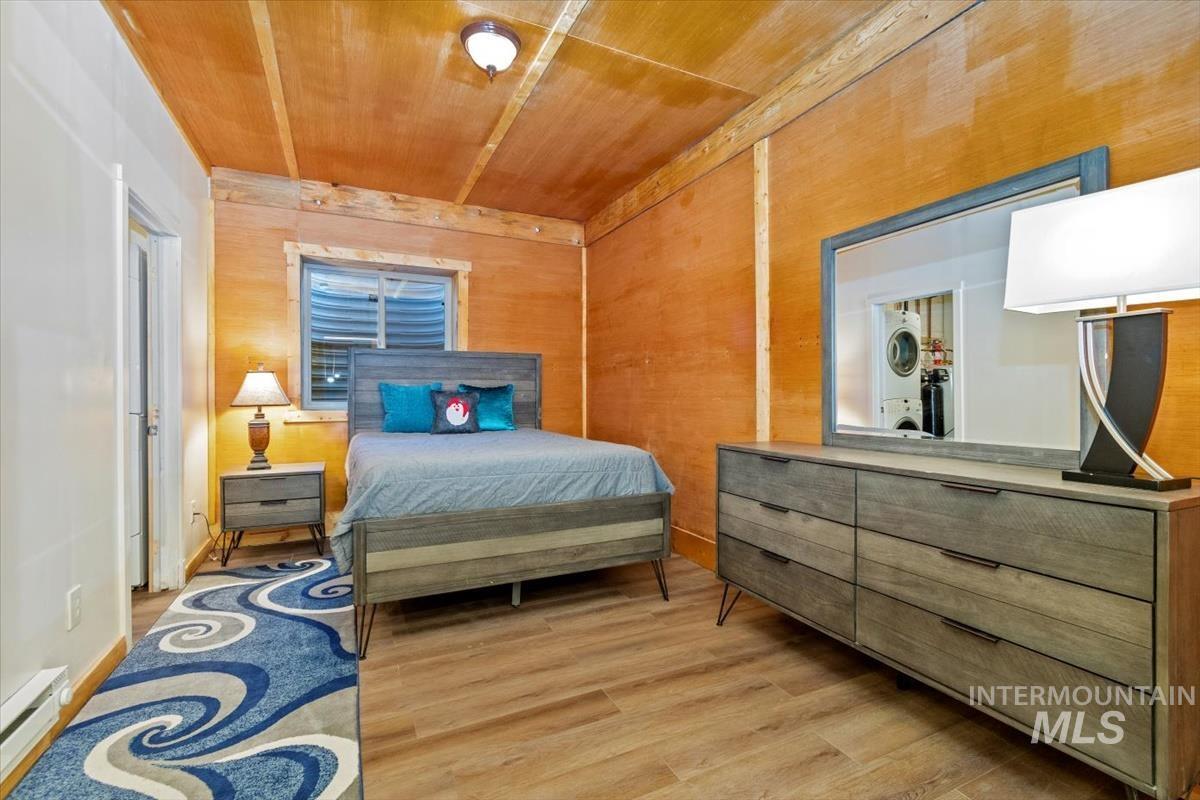 Bedroom featuring light wood-style floors, wooden ceiling, a baseboard heating unit, wood walls, and stacked washing machine and dryer