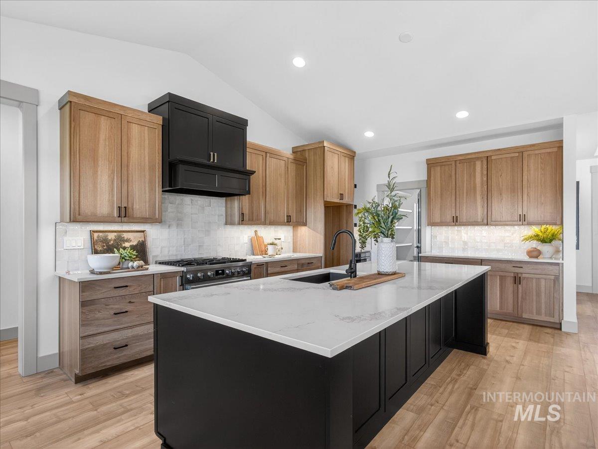 Kitchen with lofted ceiling, dark cabinets, an island with sink, light stone counters, and light wood-style floors