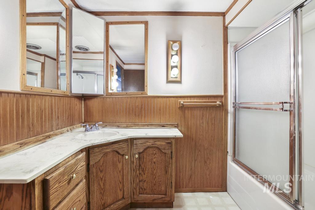 Full bathroom featuring light flooring, vanity, a wainscoted wall, bath / shower combo with glass door, and wooden walls