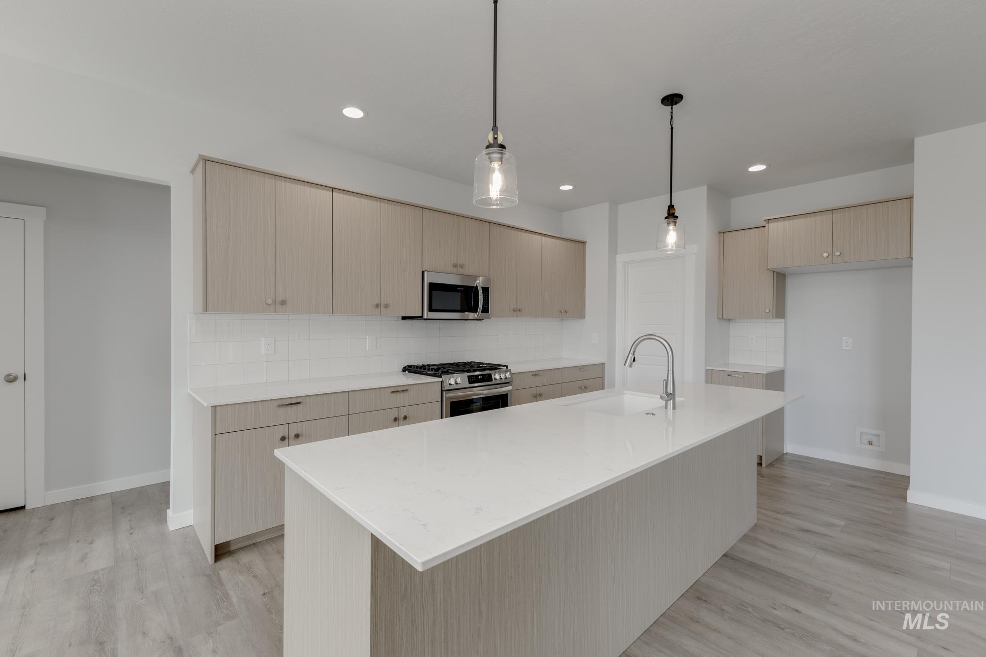 Kitchen with decorative backsplash, light stone counters, light brown cabinetry, stainless steel appliances, and a center island with sink