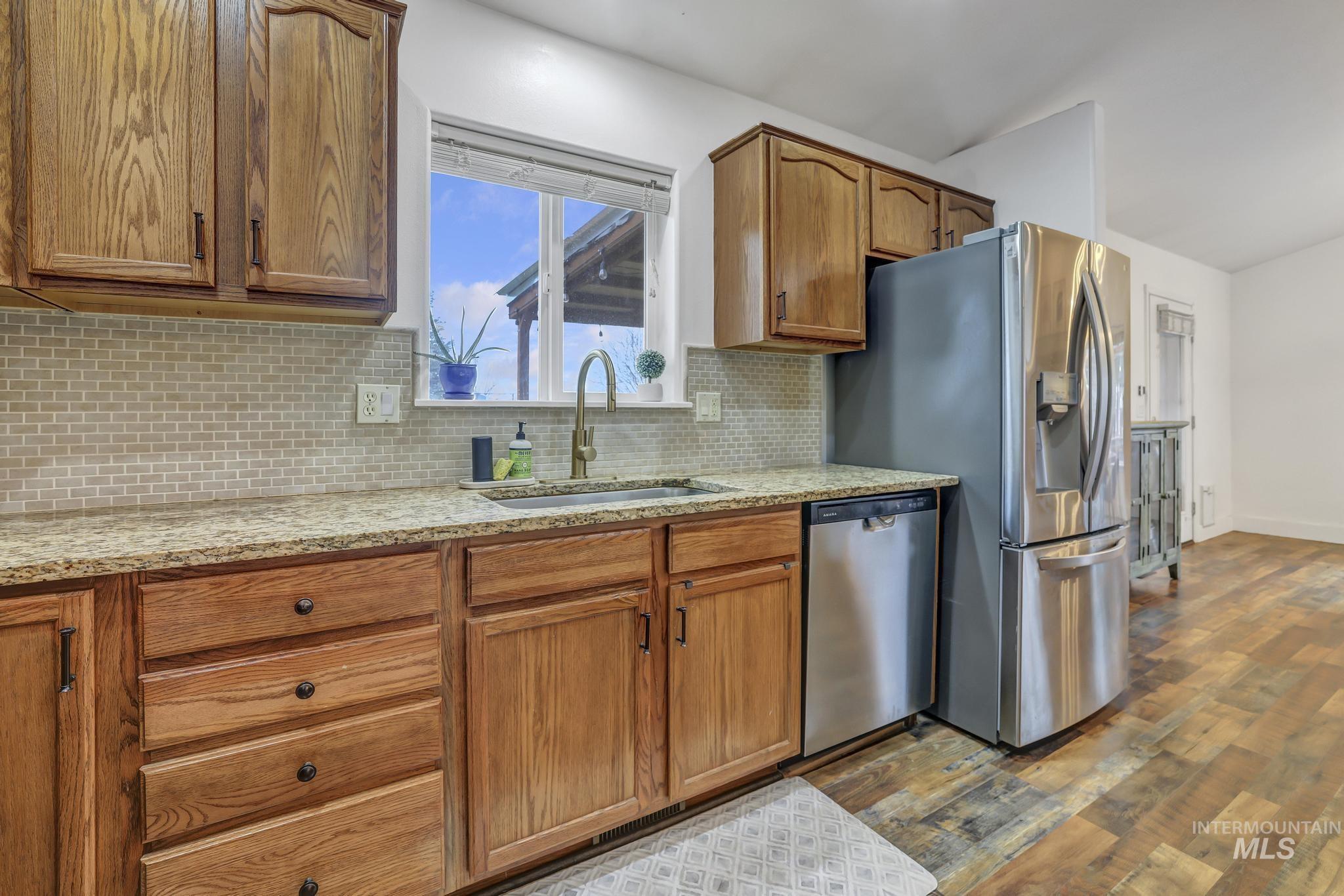 Kitchen with brown cabinetry, light stone counters, stainless steel appliances, backsplash, and dark wood-style flooring
